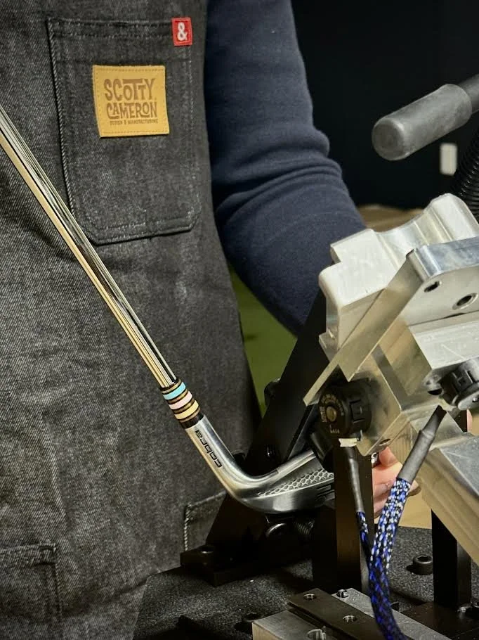 Close-up of a person working on a golf club, with a machined metal machine, possibly for club fitting or manufacturing, and a person wearing a gray vest with a 'Scotty Cameron' title patch.
