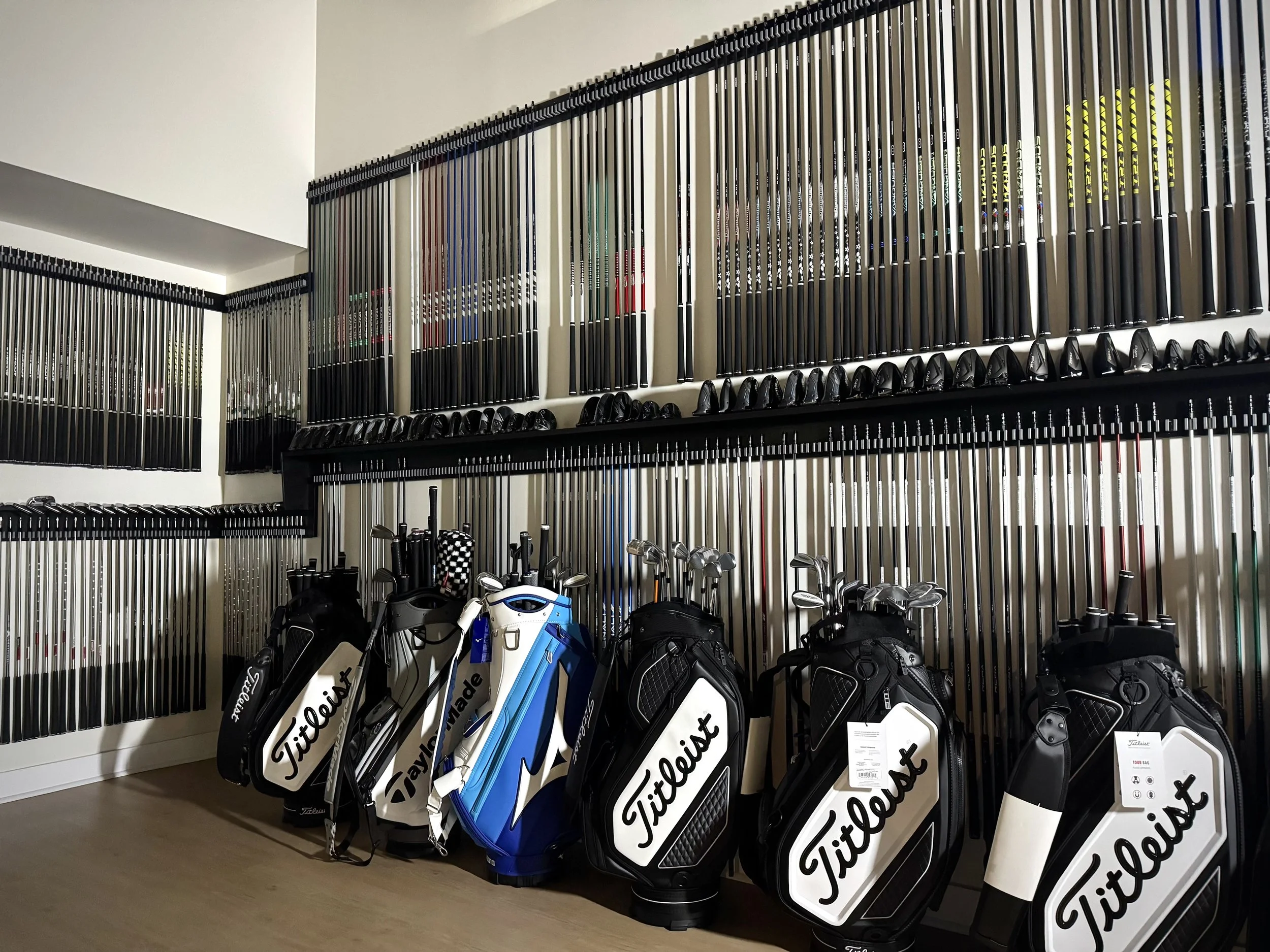 Golf clubs organized on a wall display with various brands and models. Several golf bags with brand labels are displayed on the floor in front of the wall.