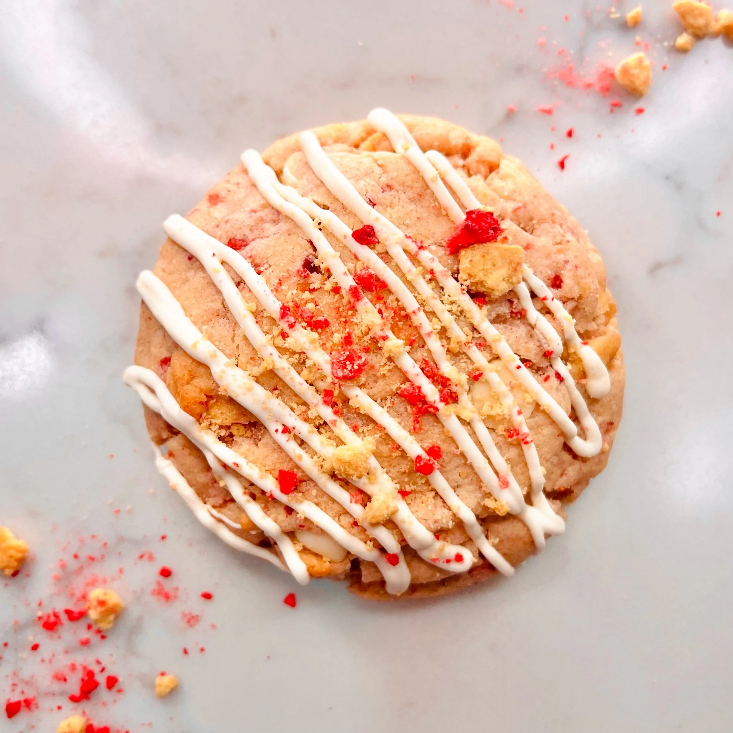 Close-up of a decorated cookie with white icing and red sprinkles, on a silver surface with crumbs and sprinkles scattered around.
