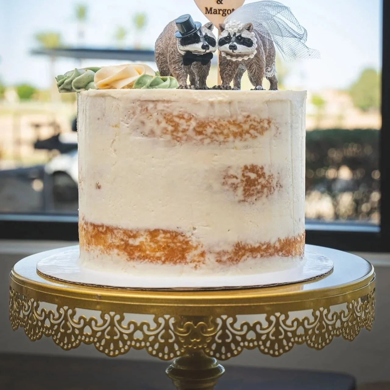 A stacked cake with semi-naked frosting and two raccoon figurines on top, dressed as a bride and groom, with a yellow cake stand.