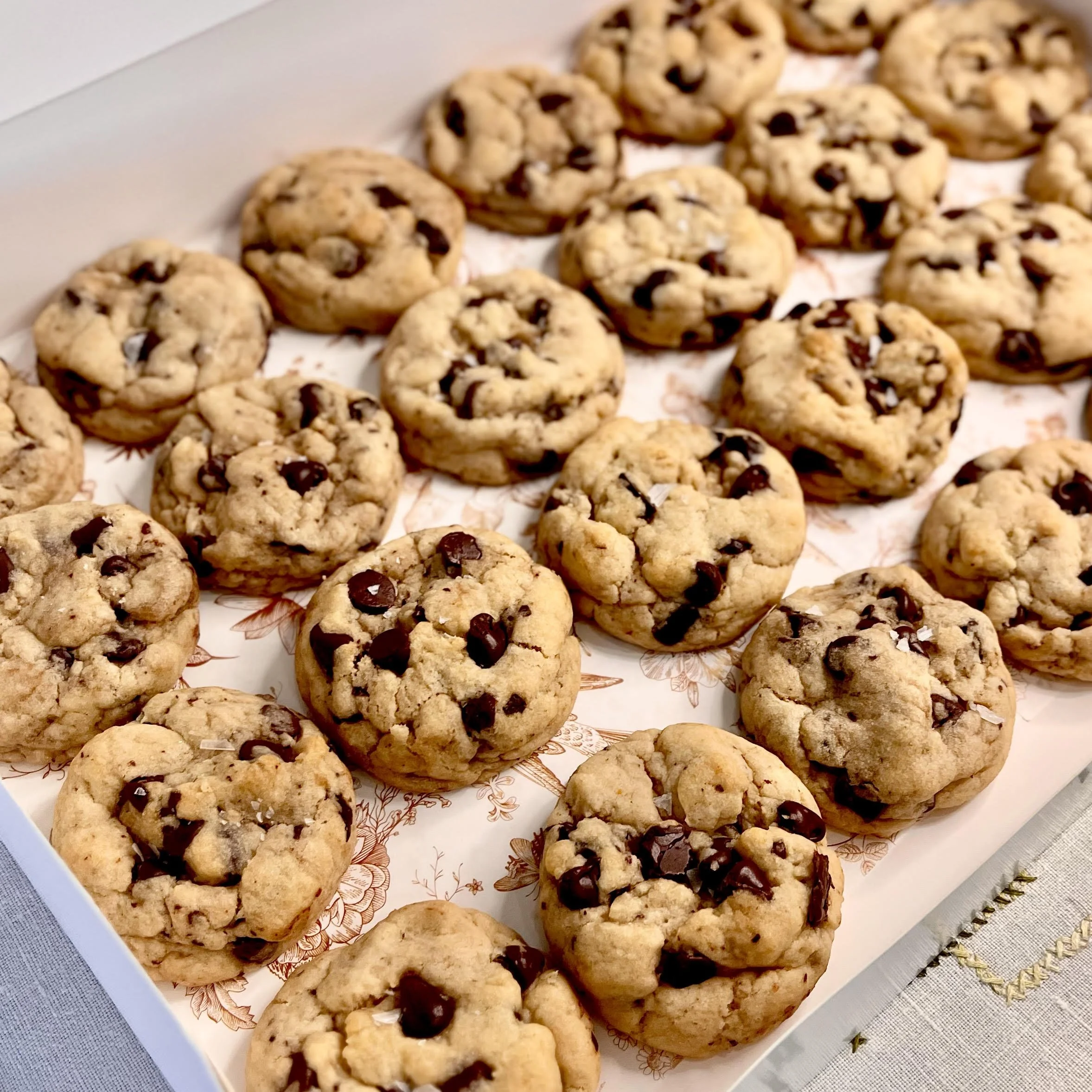 A tray of freshly baked chocolate chip cookies with some broken pieces, placed on decorative paper on a light-colored surface.