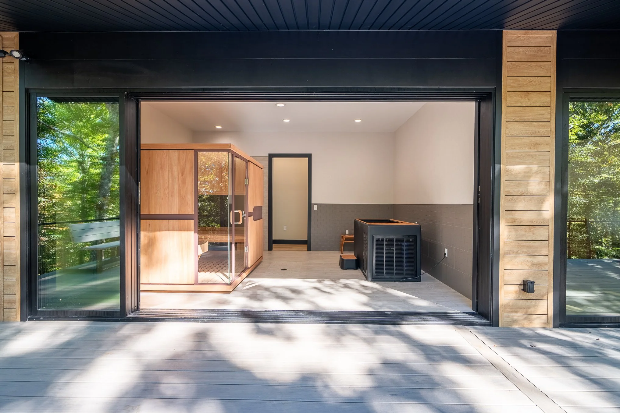 Interior view of a modern home sauna room with wooden walls and glass door, seen from a patio with wooden flooring, surrounded by trees.