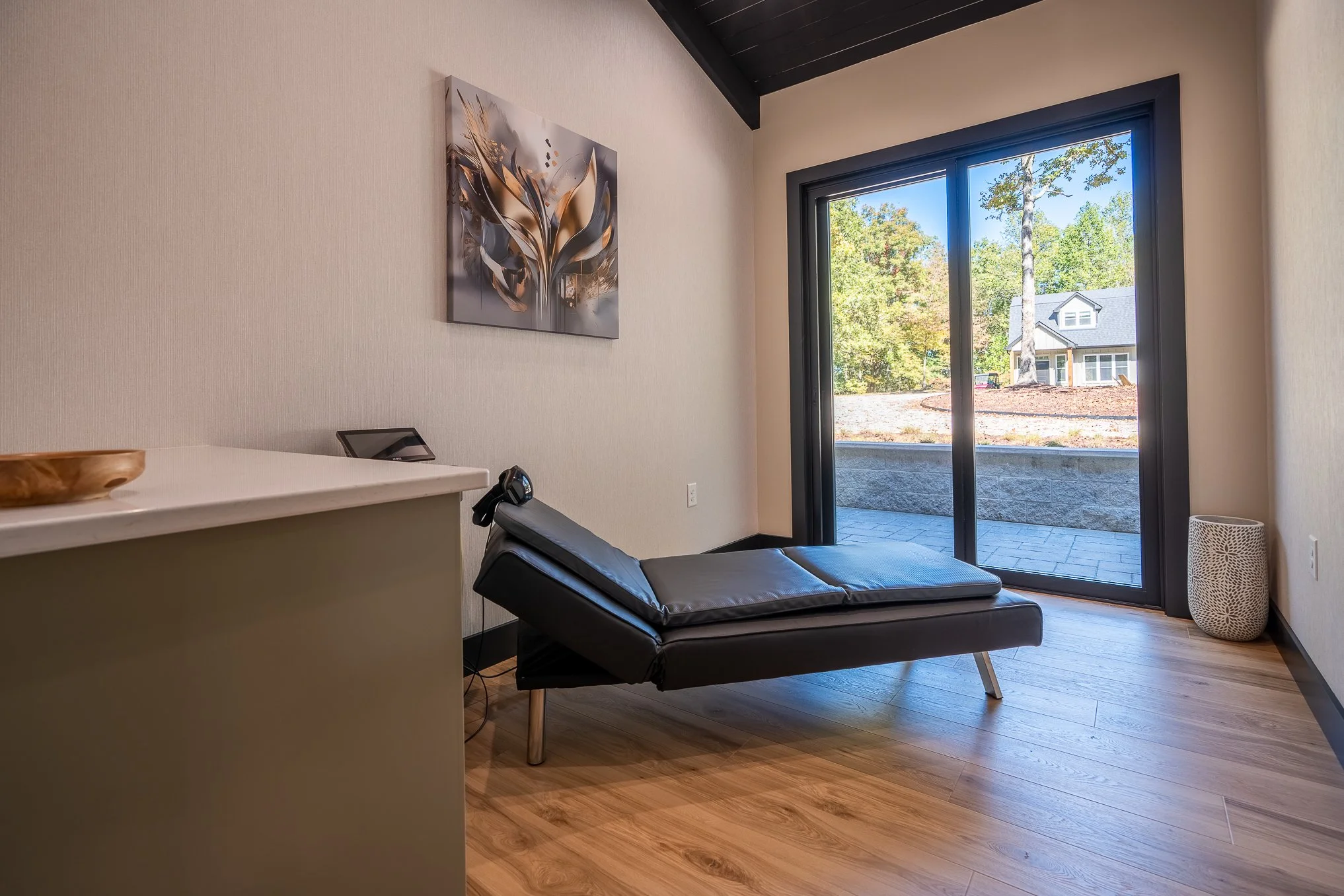 Modern interior room with black chaise lounge, abstract wall art, large glass door leading outside with trees and a house visible, wooden floor, and a patterned white side table.