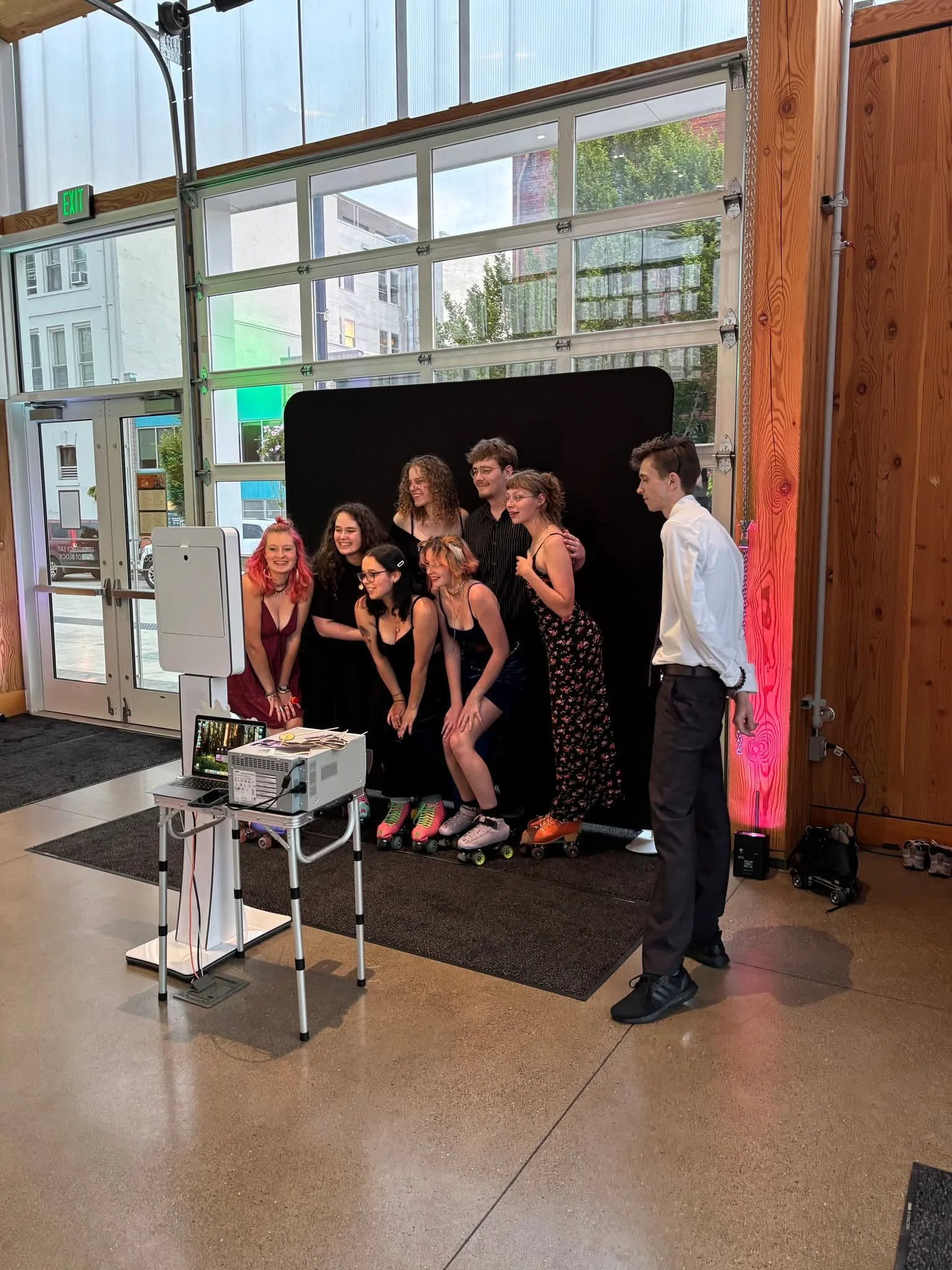 Group of diverse women and a man standing outside near a white wall with potted flowers, posing in a photobooth with a blank white photo booth prop, smiling and showing playful expressions.