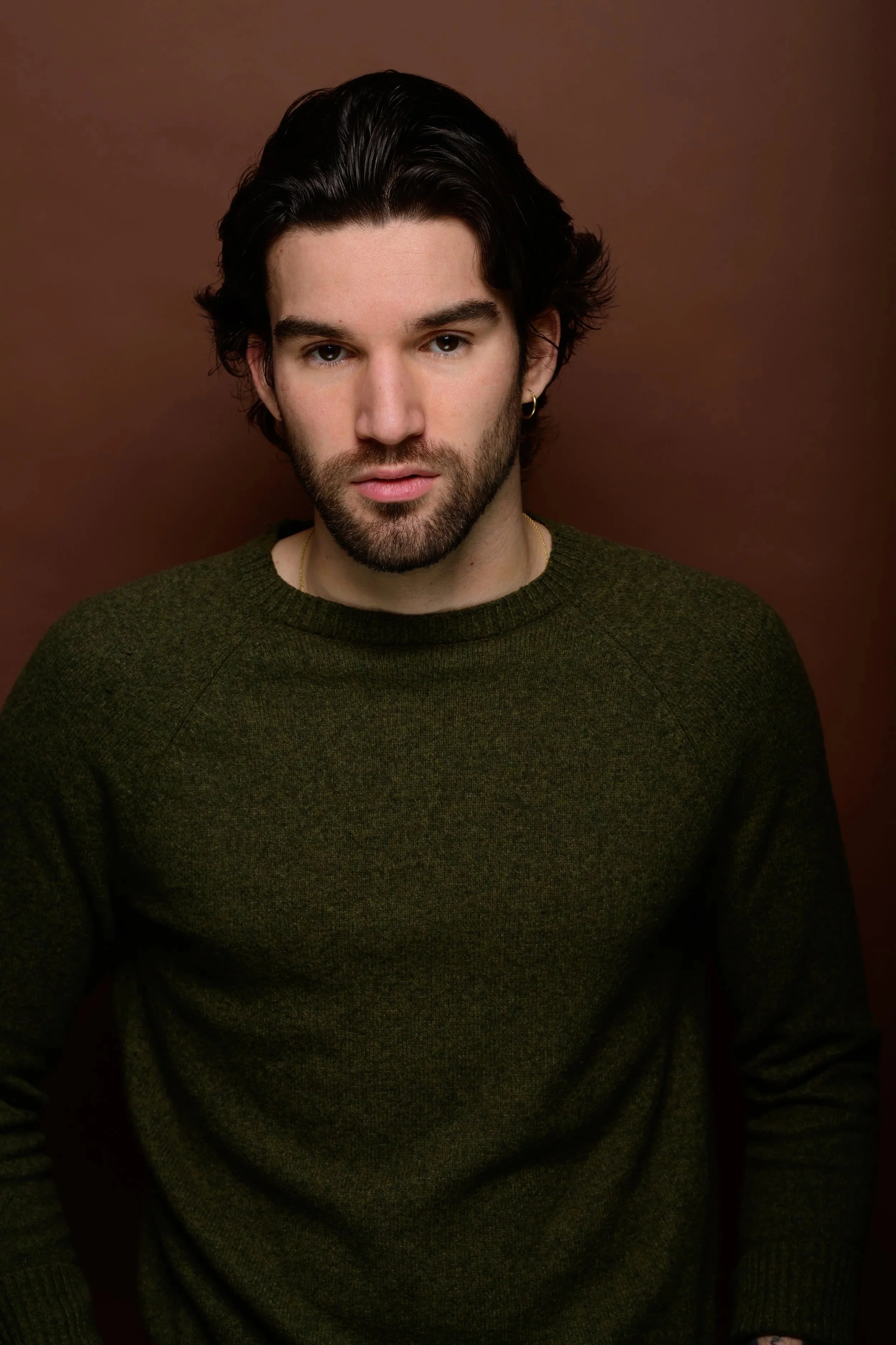 A young man with dark, wavy hair and a beard, wearing a dark green sweater, gazing at the camera against a brown background.