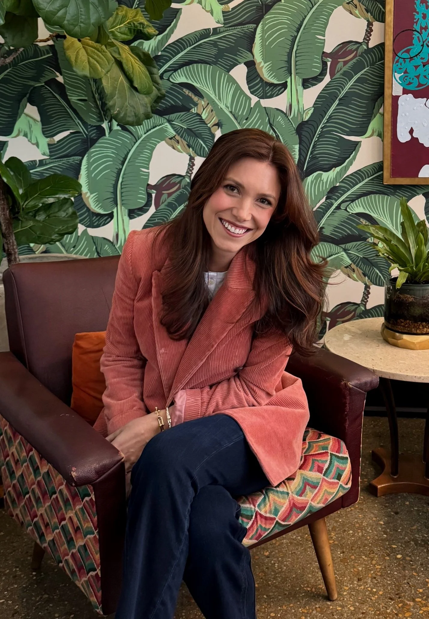 A woman sitting on a colorful patterned chair, smiling, wearing a pink blazer and dark jeans, in a room with tropical-themed wallpaper and green plants.