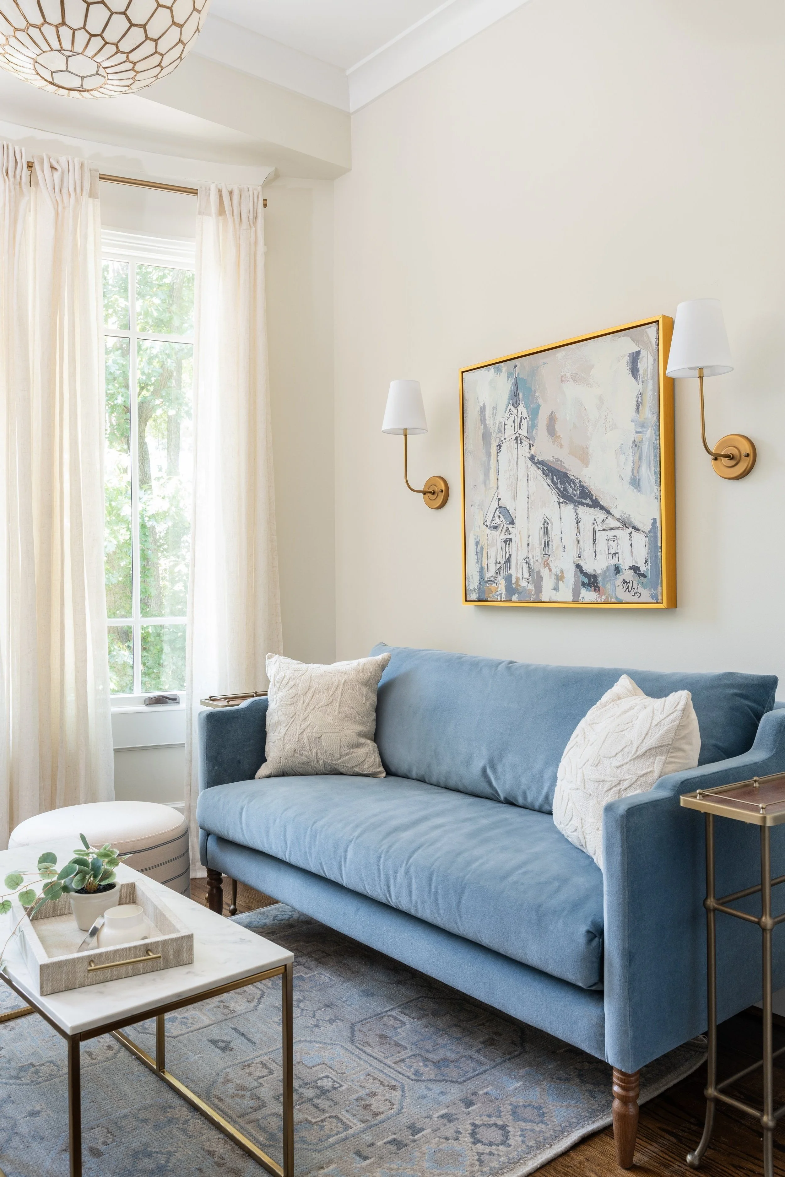 A cozy living room with a light blue sofa, two white textured pillows, a marble-top side table with a plant, a patterned rug, cream curtains, and a large window showing trees outside. A painting of a church and two wall-mounted lamps are on the wall above the sofa.