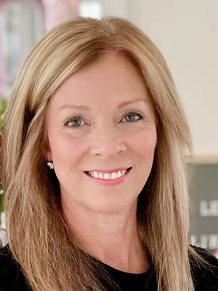 Close-up of a smiling woman with long blonde hair, wearing earrings, in an indoor setting.