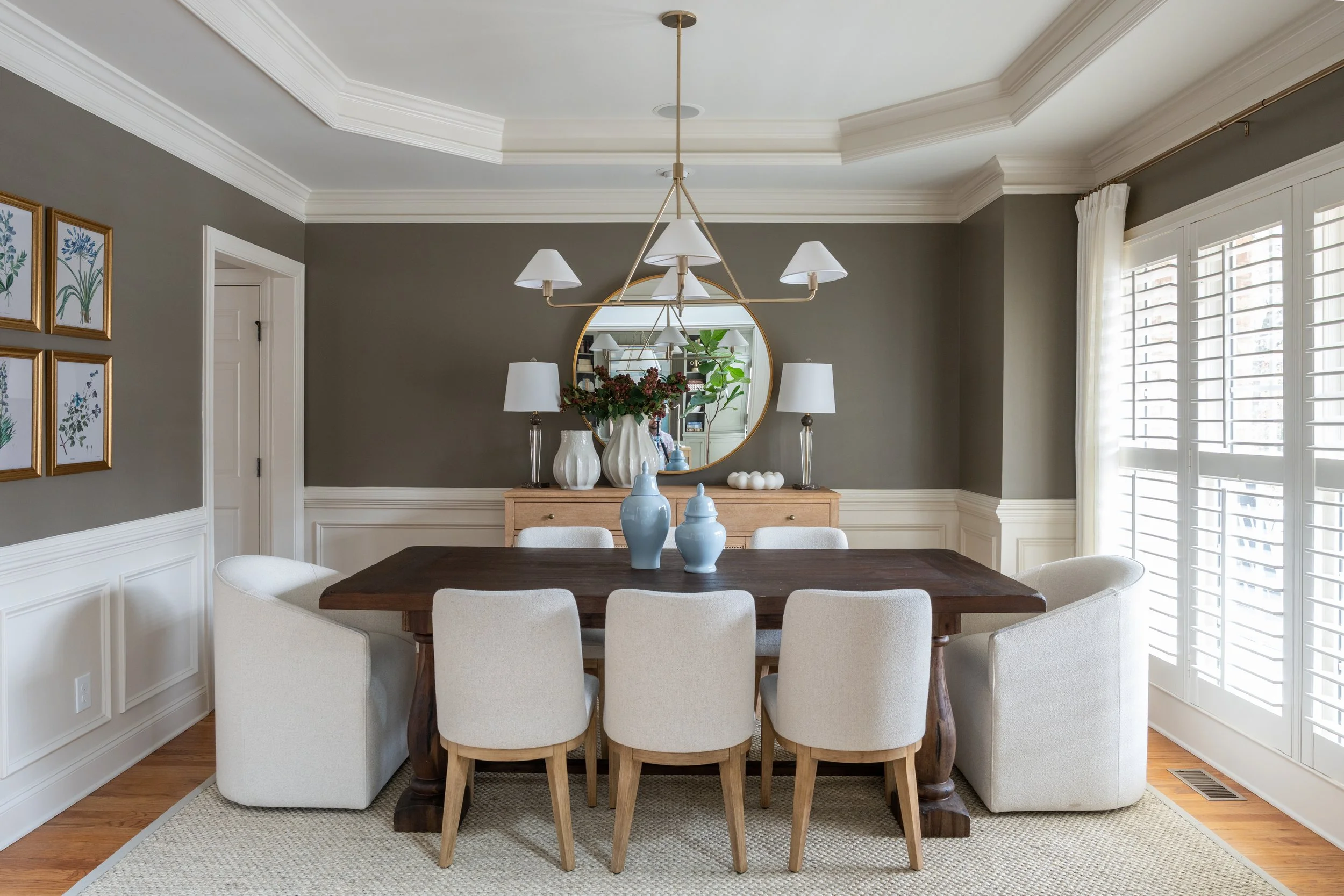 A dining room with a large wooden table surrounded by six white upholstered chairs and two armchairs, a wooden sideboard with vases, candles, and a round mirror, a chandelier, wall art, and windows with white shutters.