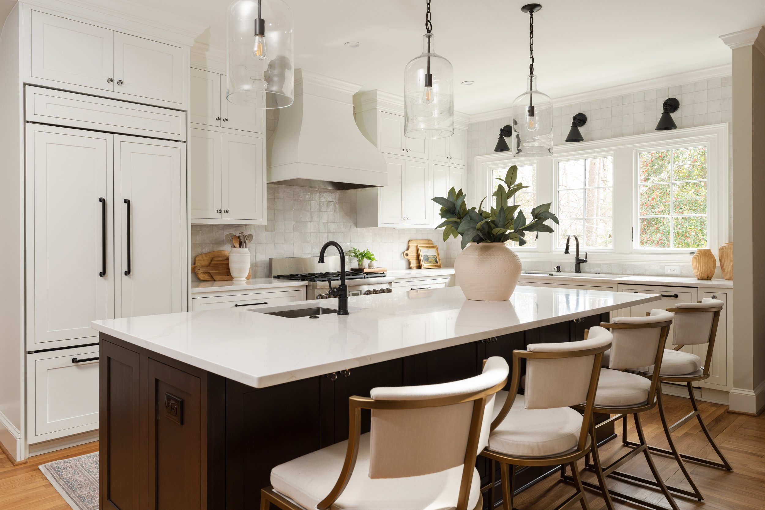 An elevated, traditional kitchen with off-white cabinets, black hardware, a central island with a white countertop, a vase with green leaves, pendant lights, and a window with a view of trees.