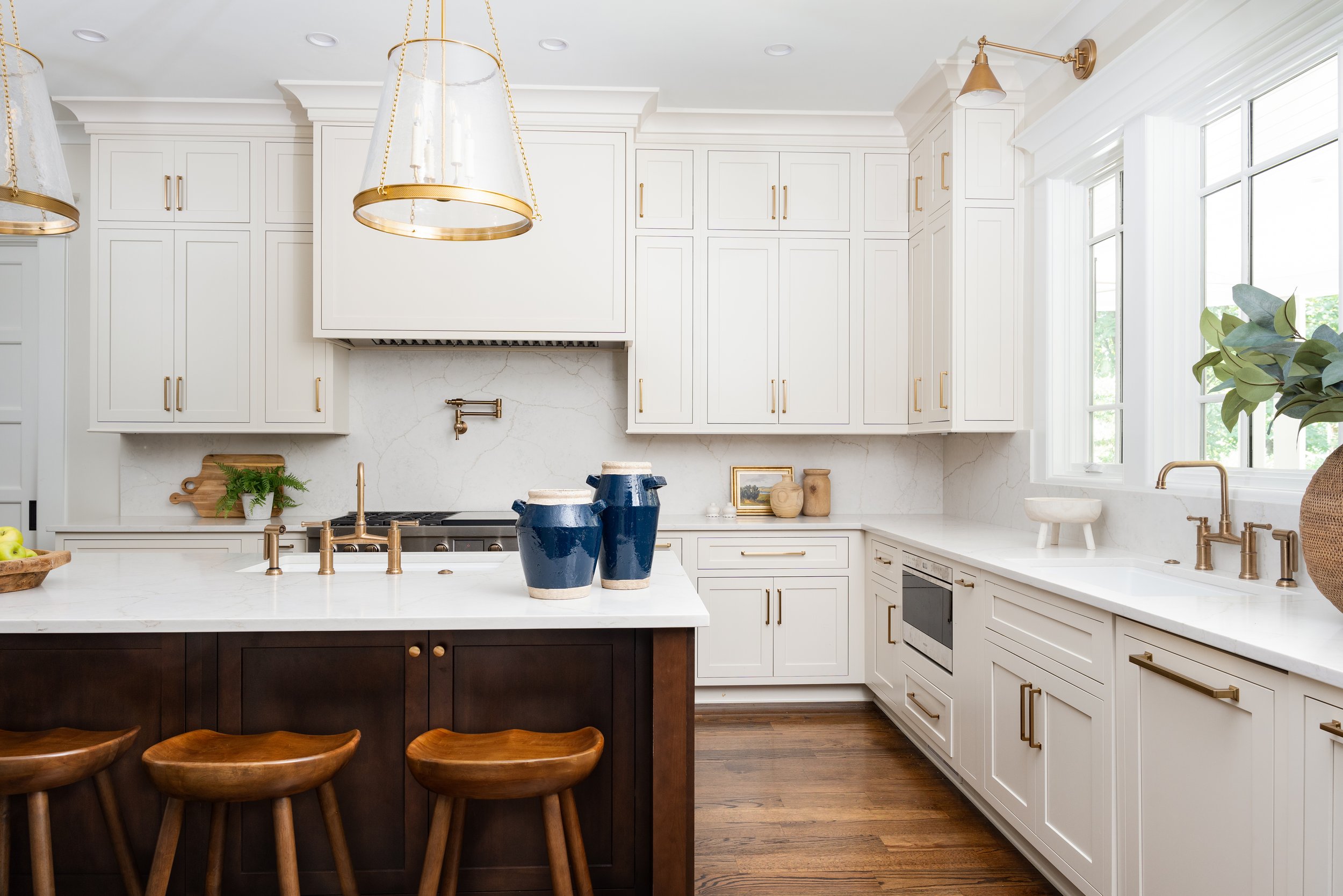 Bright modern kitchen with white cabinets, marble countertops, wooden flooring, and a dark wood island with wooden stools. Accents include gold hardware, lighting fixtures, blue and white ceramic jars, and green plants near a large window.