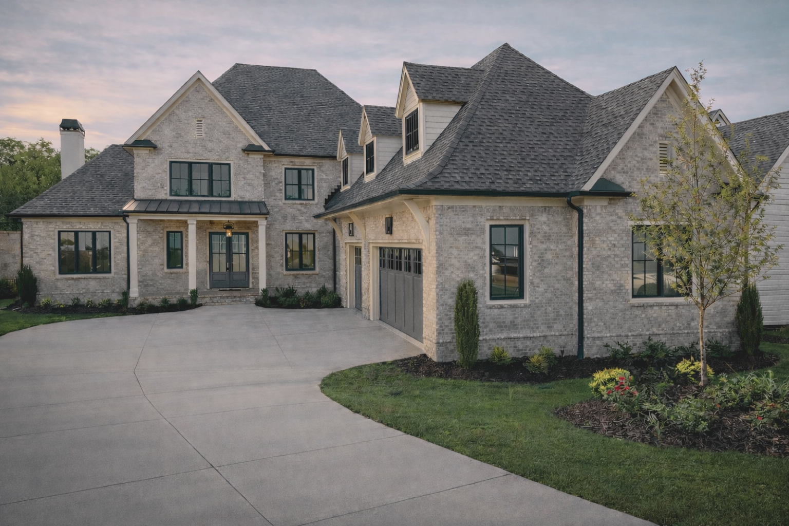 Large modern house in Milton, GA with light brick exterior, multiple gabled roofs, black window frames, a concrete driveway, and landscaped lawn with young trees and shrubs. New Construction Home.