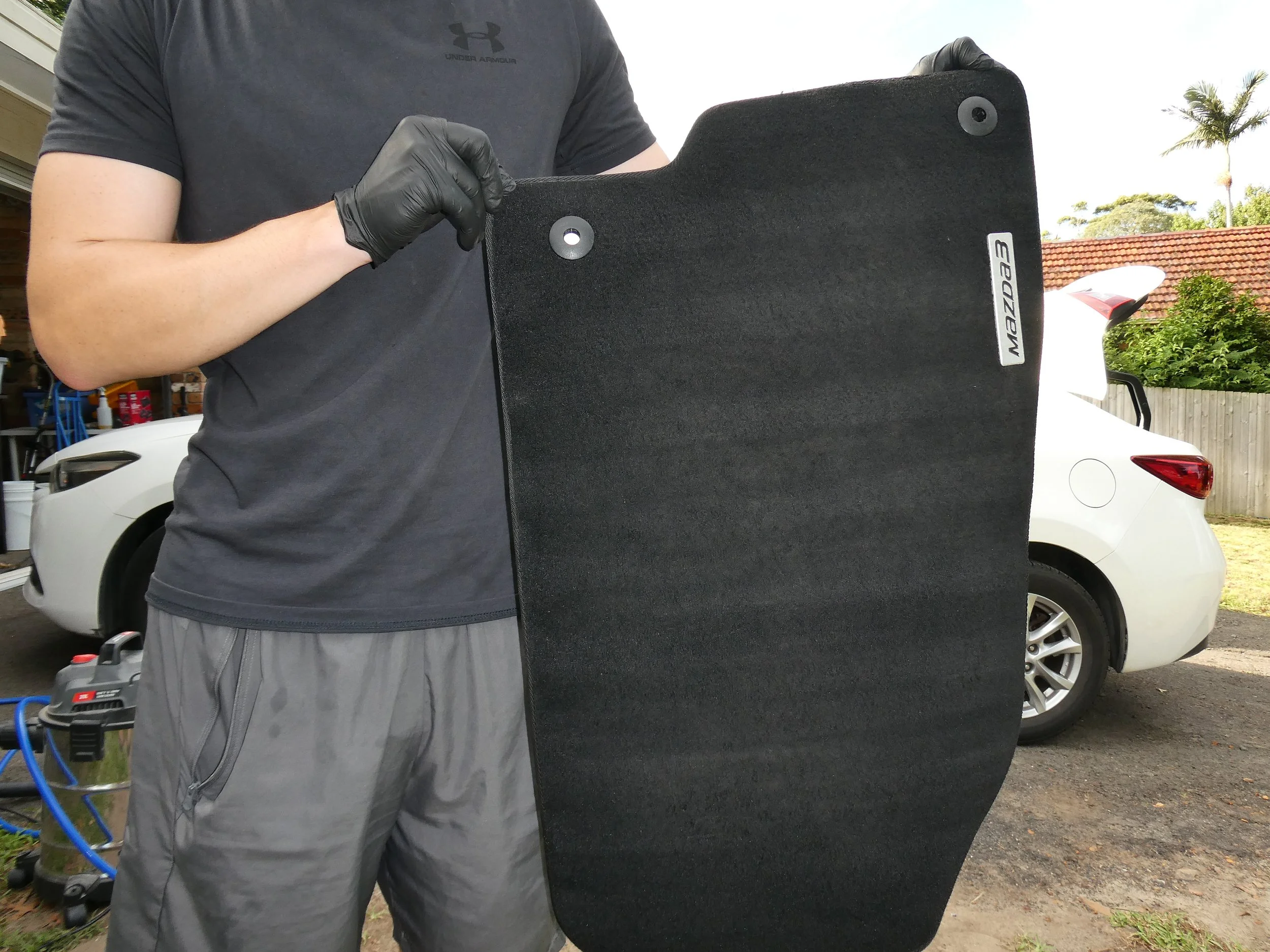 A person wearing a black t-shirt and gray shorts holding a black portable car windshield shield, with a white Mazda car and a residential backyard with trees and a red-tiled roof in the background.
