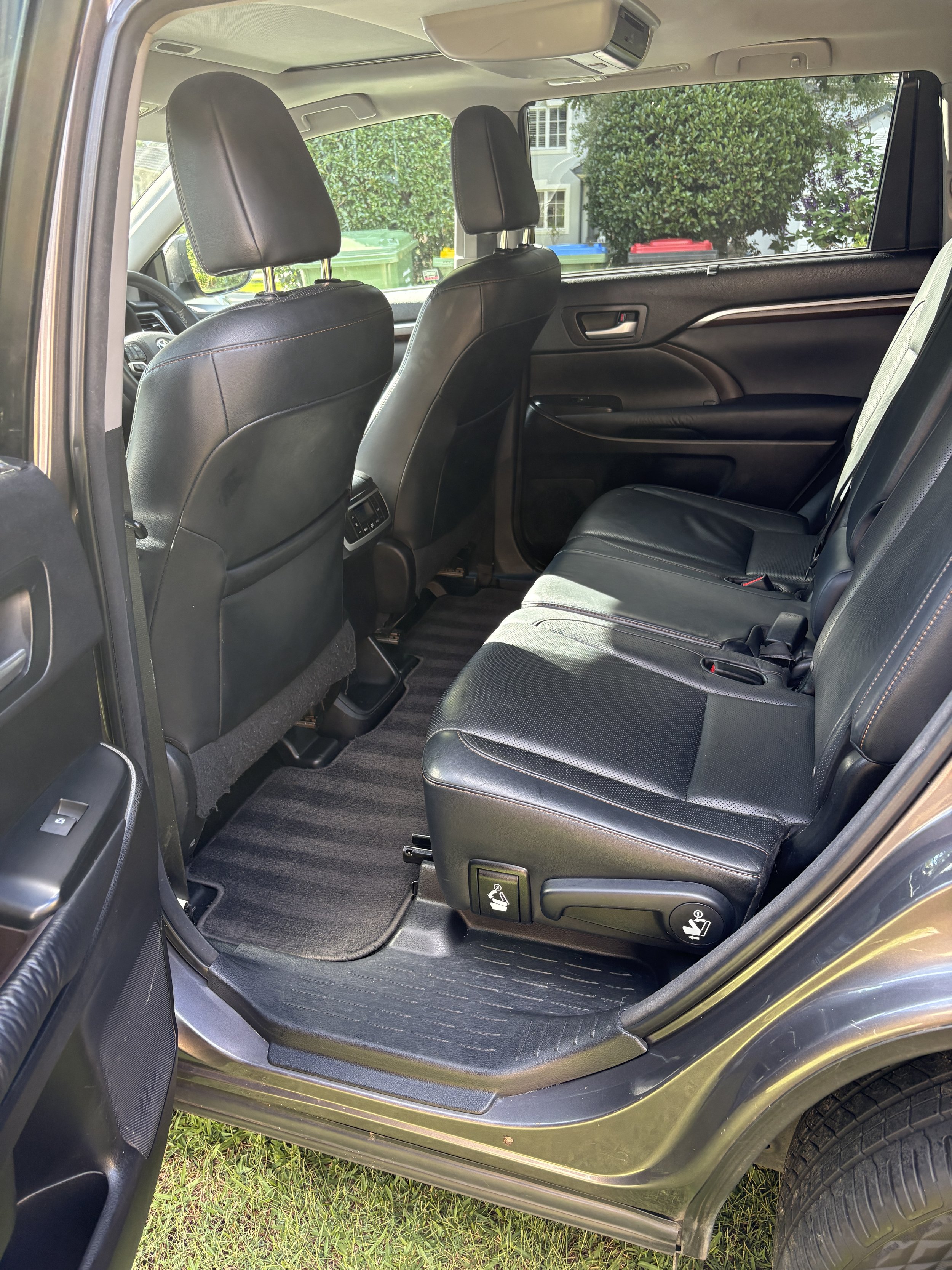 Interior view of a car's back seat area with black leather seats, black floor mats, and headrests, with a window showing green bushes and some houses outside.