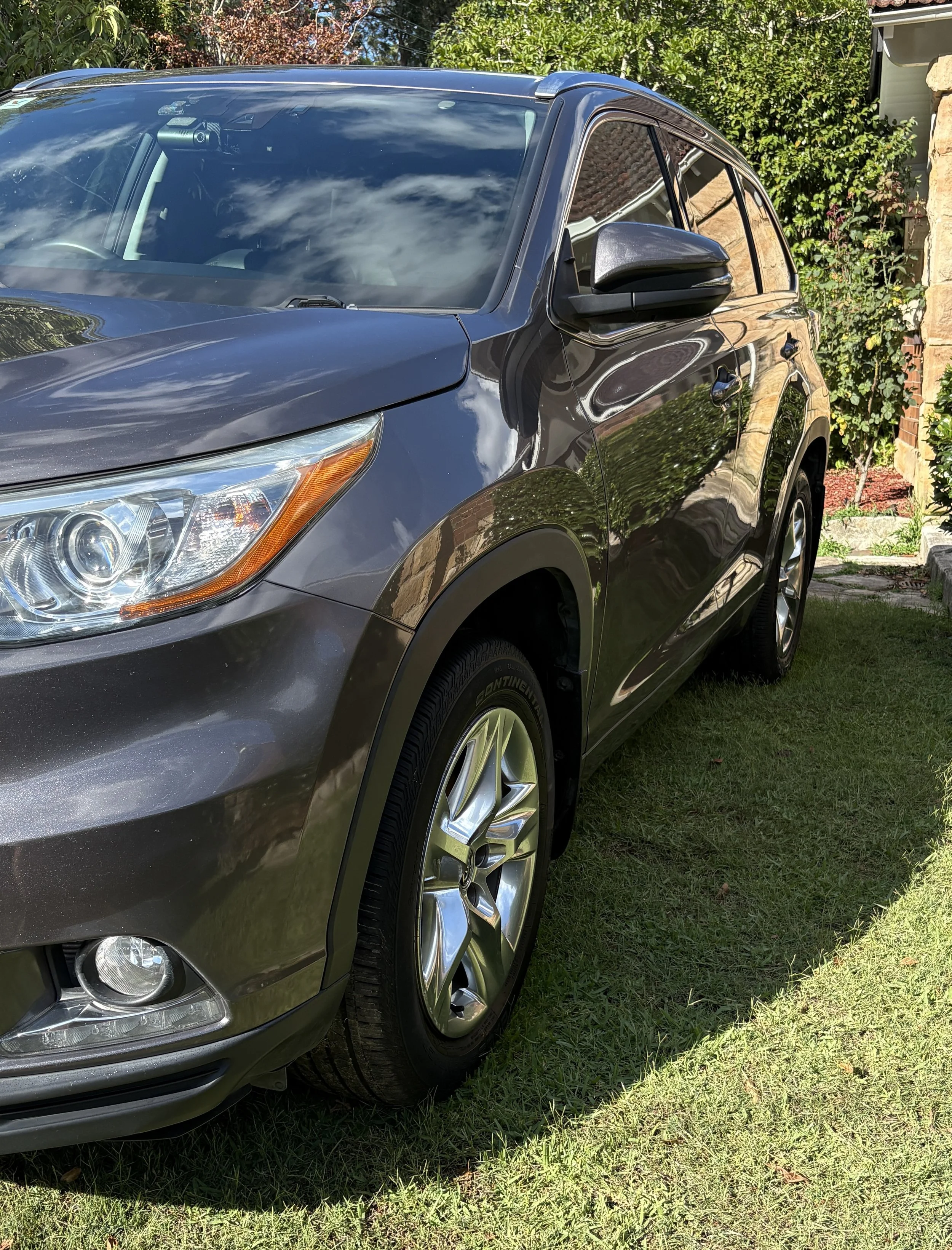 Front and side view of a dark gray SUV parked on a grassy yard with bushes and trees in the background, reflecting the sky on its windshield and body.