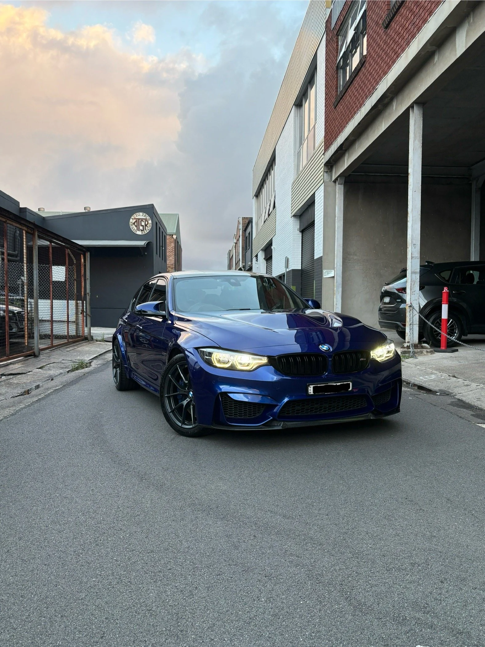 A blue BMW sports car parked on a city street surrounded by modern buildings, with bright headlights and black rims.
