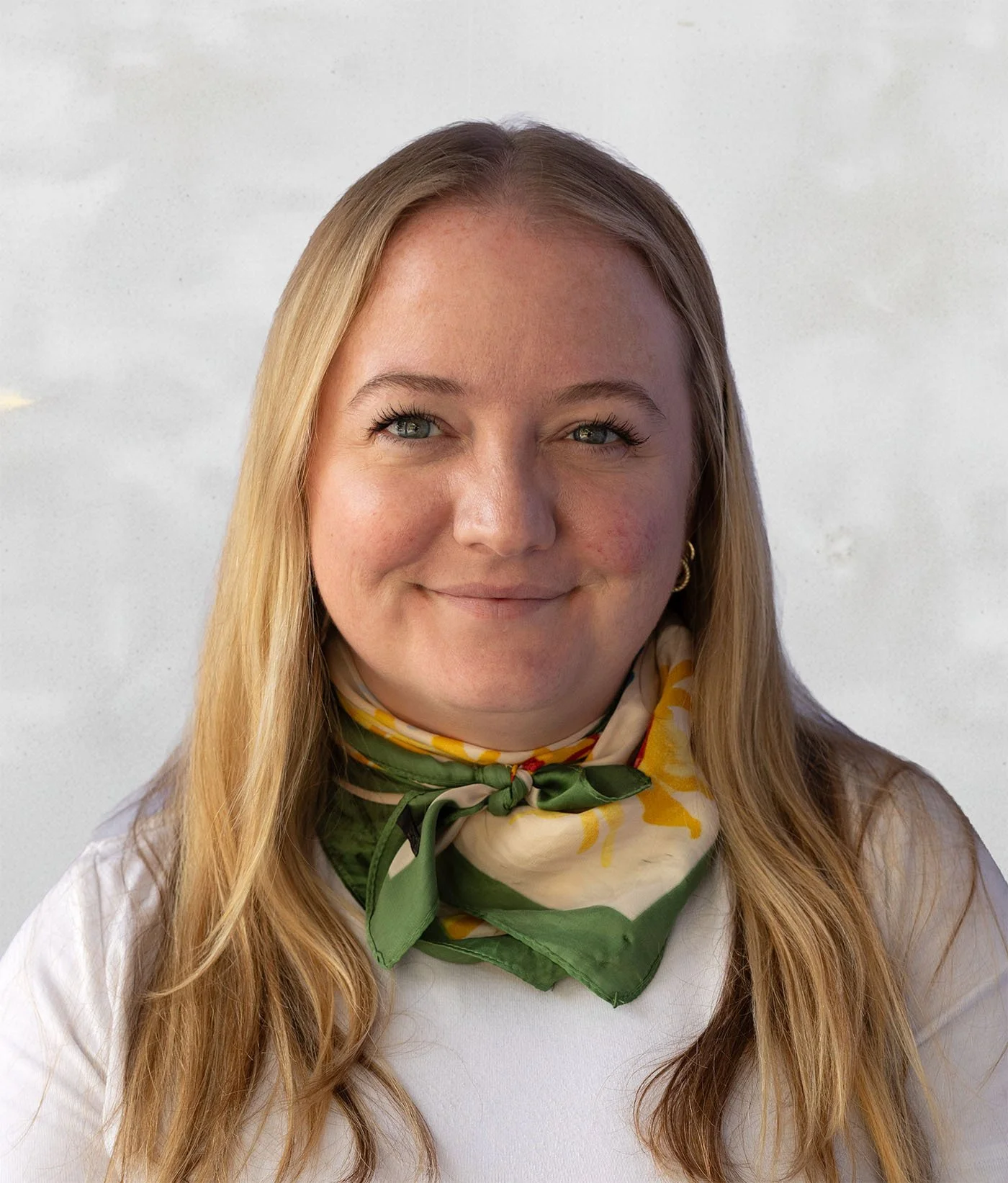 A woman with long blonde hair smiling, wearing a white top and a green and yellow patterned scarf around her neck, against a plain background.