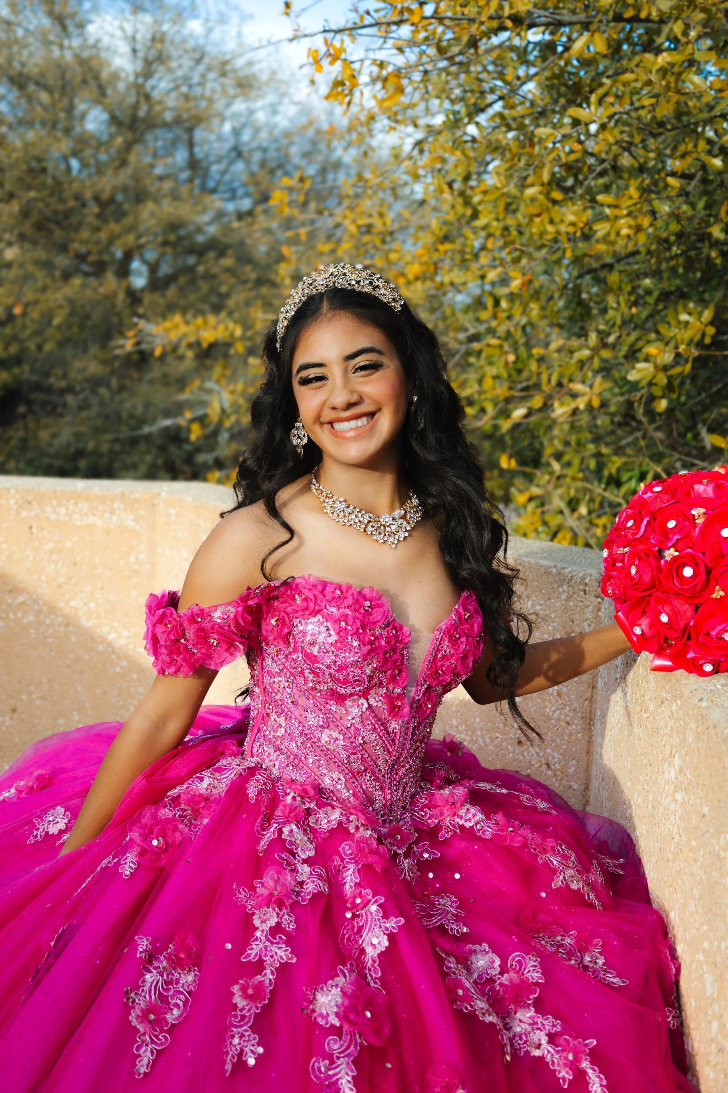 Young woman in a vibrant pink, ornate gown with floral embroidery, holding a bouquet of pink roses, smiling outdoors with autumn trees in the background.
