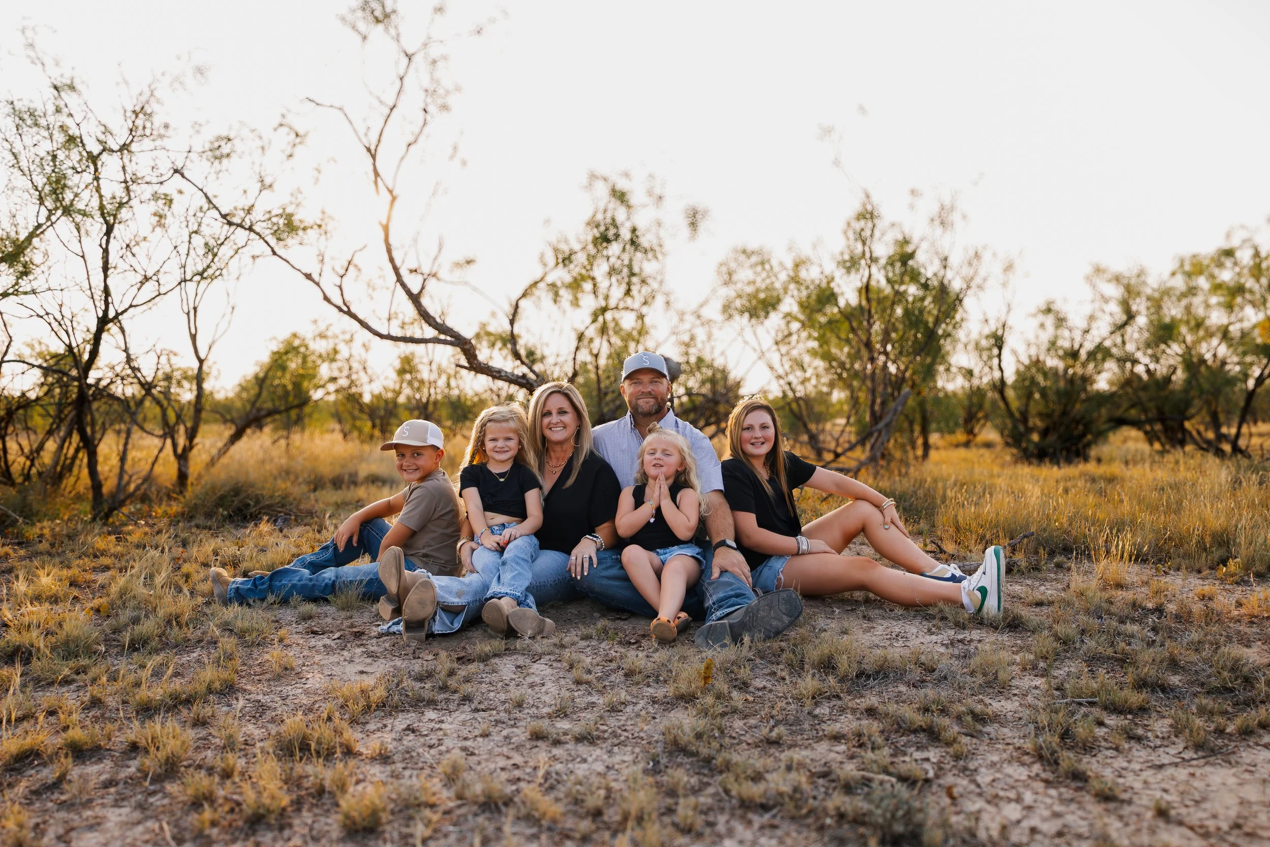 Family of six sitting on the ground outdoors in a field with trees, during sunset, smiling at the camera.