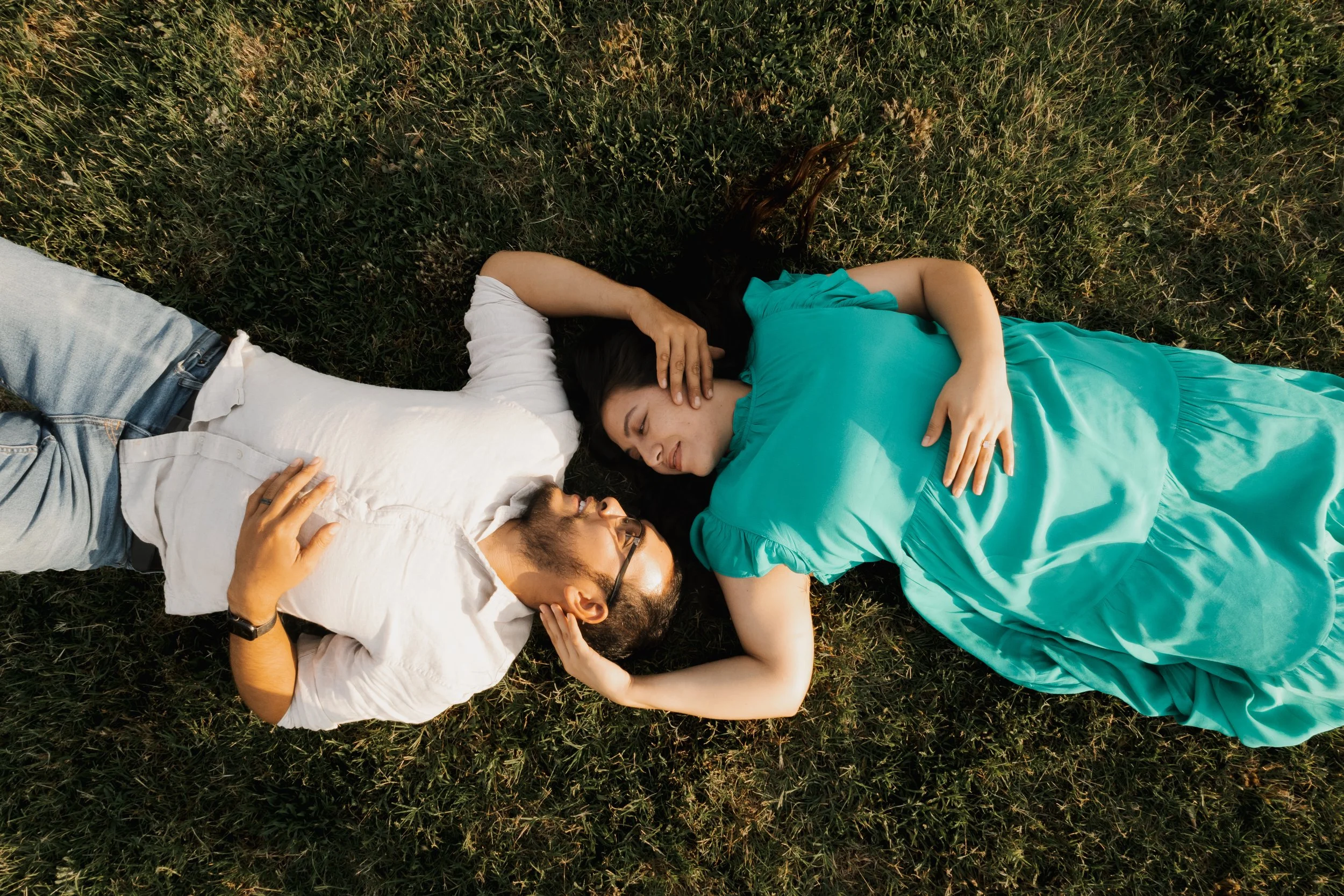 A man and a woman lying on grass, looking at each other and smiling, with the woman resting her head on her hand.