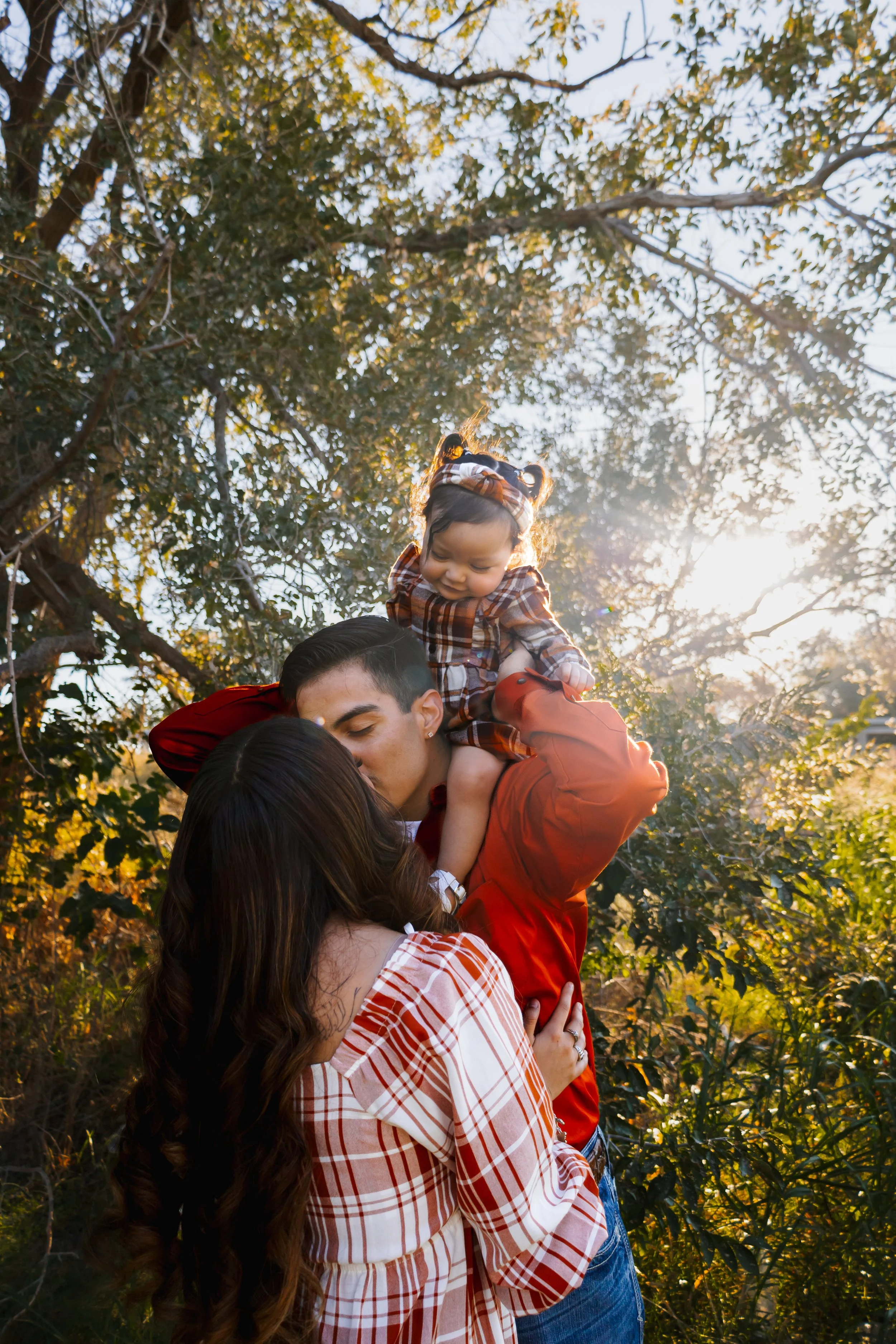 A family of three outdoors in a wooded area during the golden hour, with the sun shining brightly behind the trees. The man is holding a young girl on his shoulders, and the woman is standing close, touching his back as they share a kiss.