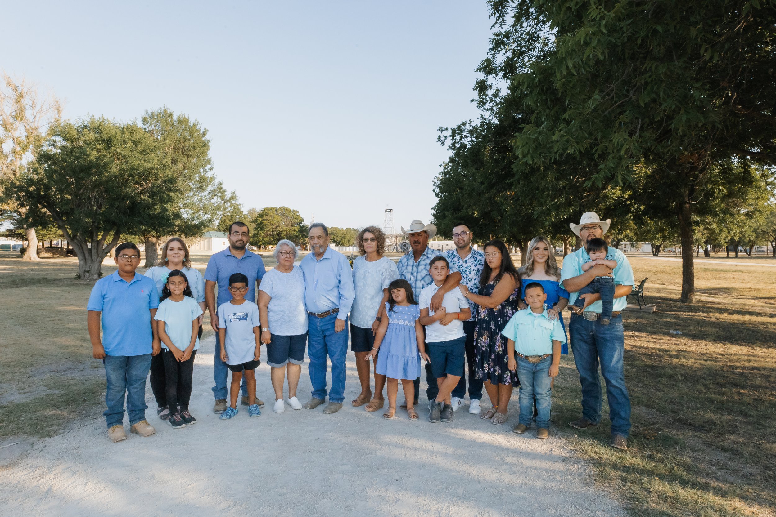 A large family group standing outdoors on a dirt path in a park with trees and open space behind them, dressed in casual summer clothing.
