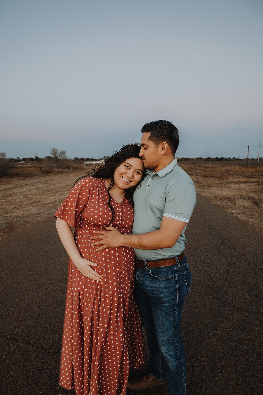 A smiling pregnant woman in a red polka dot dress holding her belly, with a man in a light blue polo shirt and jeans embracing her and kissing her forehead, standing on a dirt road in a rural area during sunset.