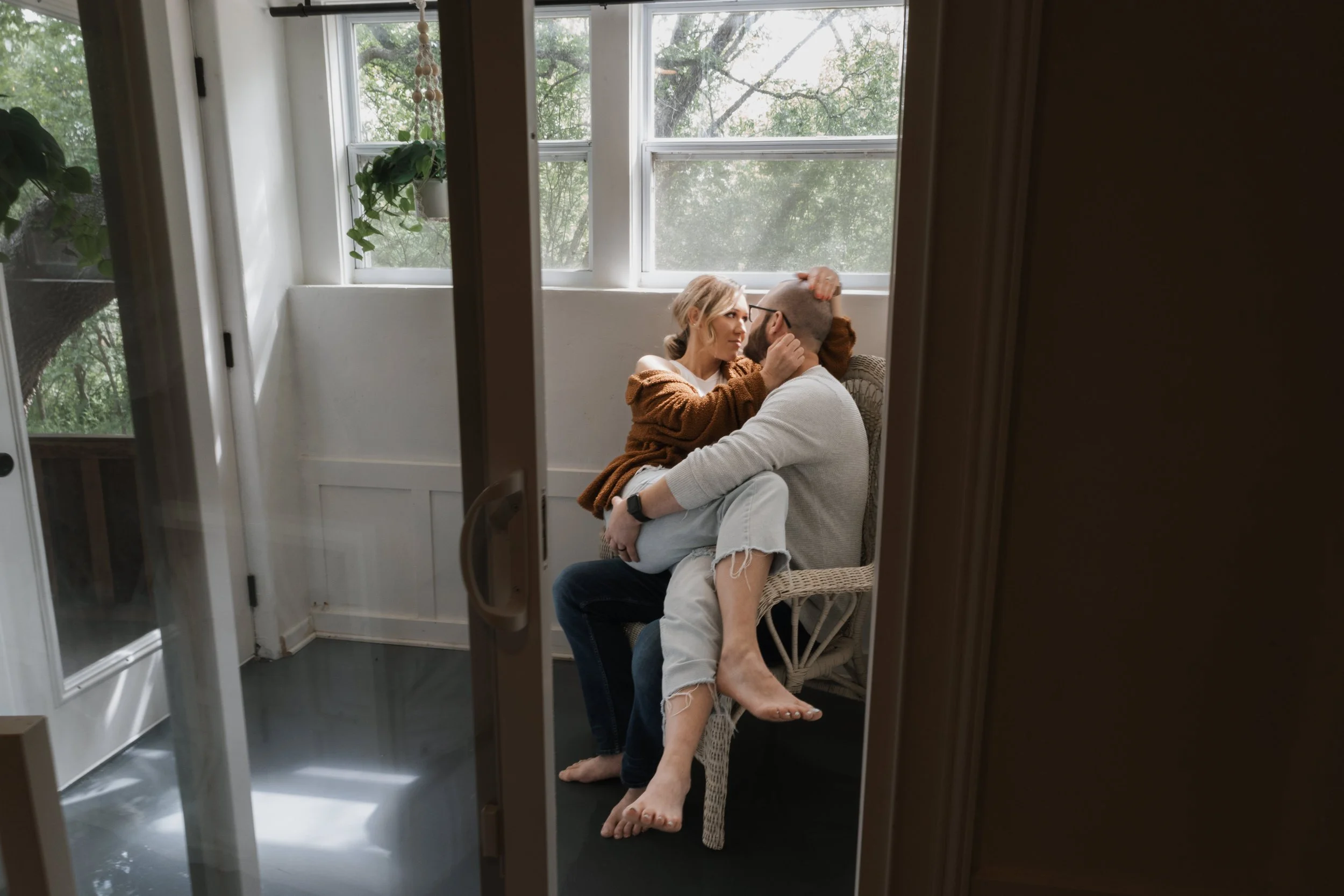 A couple sitting in a cozy sunlit room, sharing an intimate moment. The woman is sitting on the man's lap, gently touching his head, while the man holds her around the waist. They are near a large window with greenery outside.