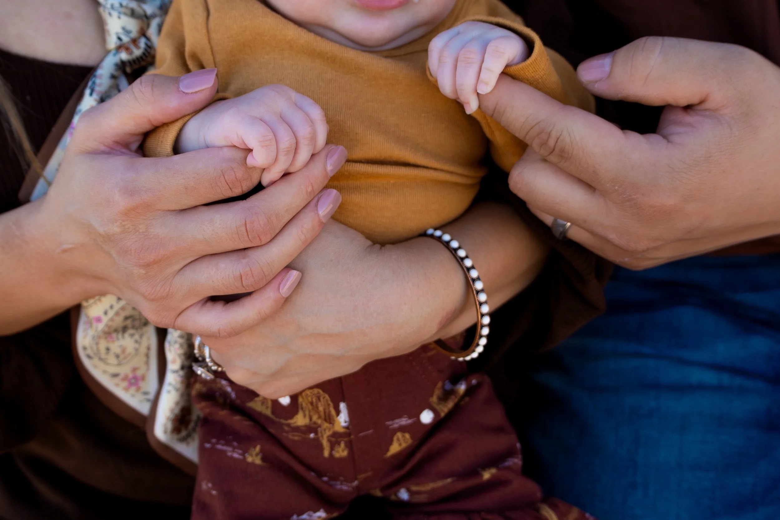 Close-up of a baby in a mustard yellow shirt with hands of two adults gently holding the baby's hands, one adult wearing a ring and the other a bracelet.