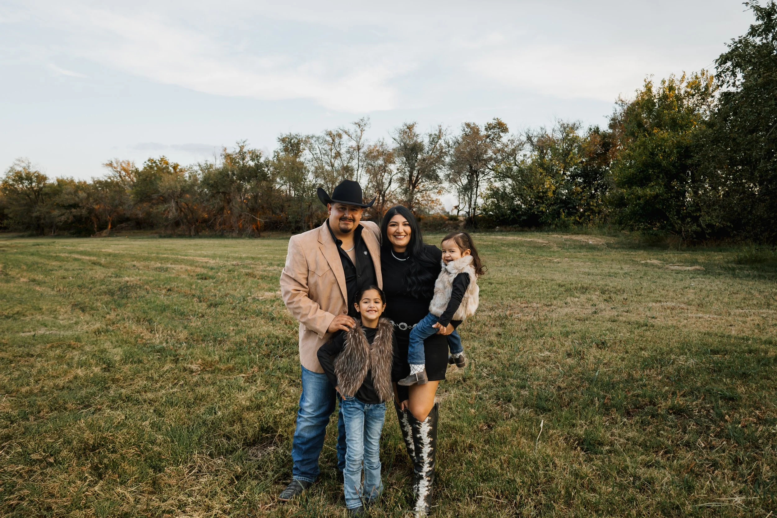 A family of four standing outdoors on a grassy field with trees in the background, smiling at the camera. The father wears a cowboy hat and beige jacket, the mother wears a black dress and patterned boots, and the two children are dressed in casual clothes with faux fur vests.