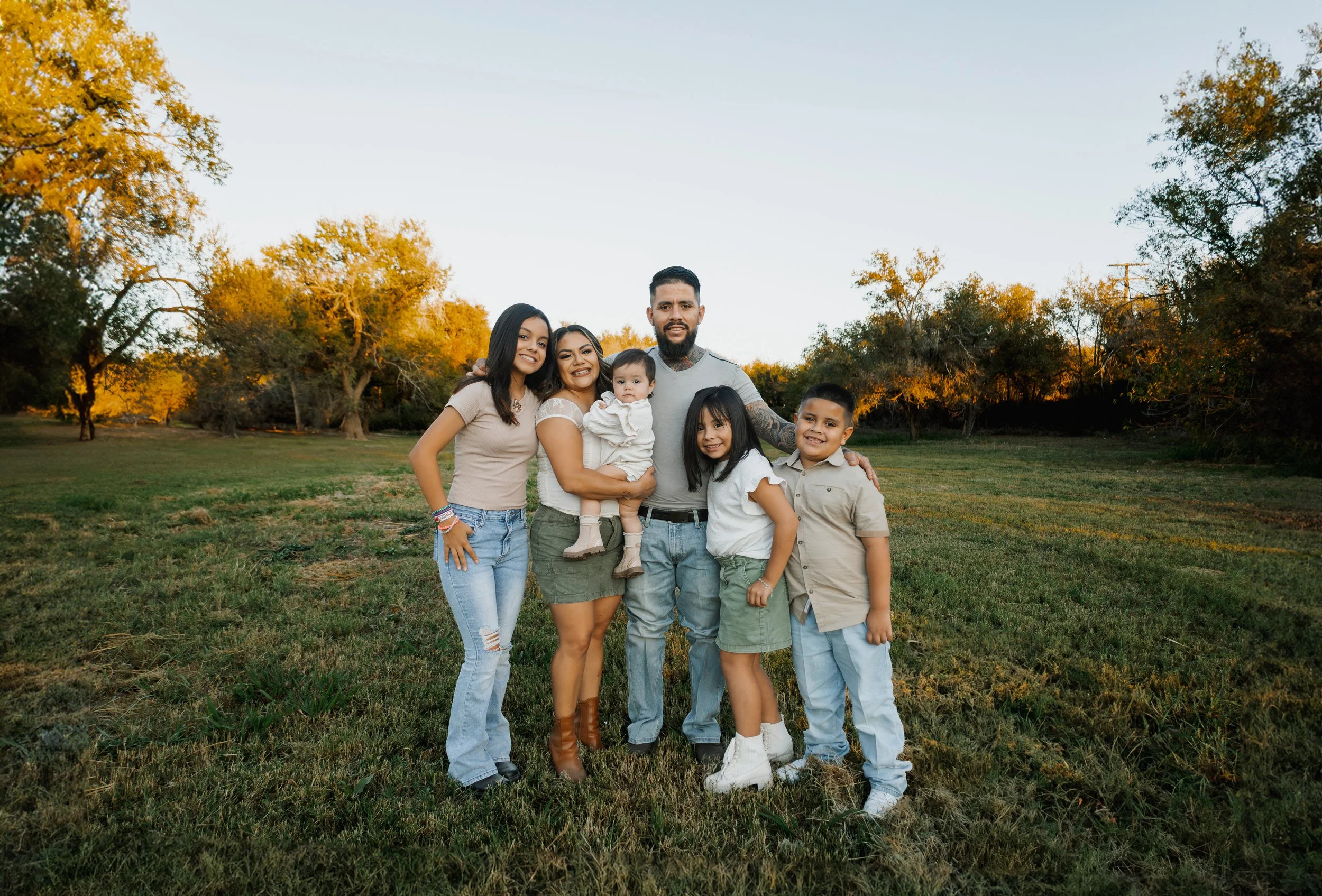 A family of six standing together outdoors in a grassy park at sunset, smiling and posing for a photo.