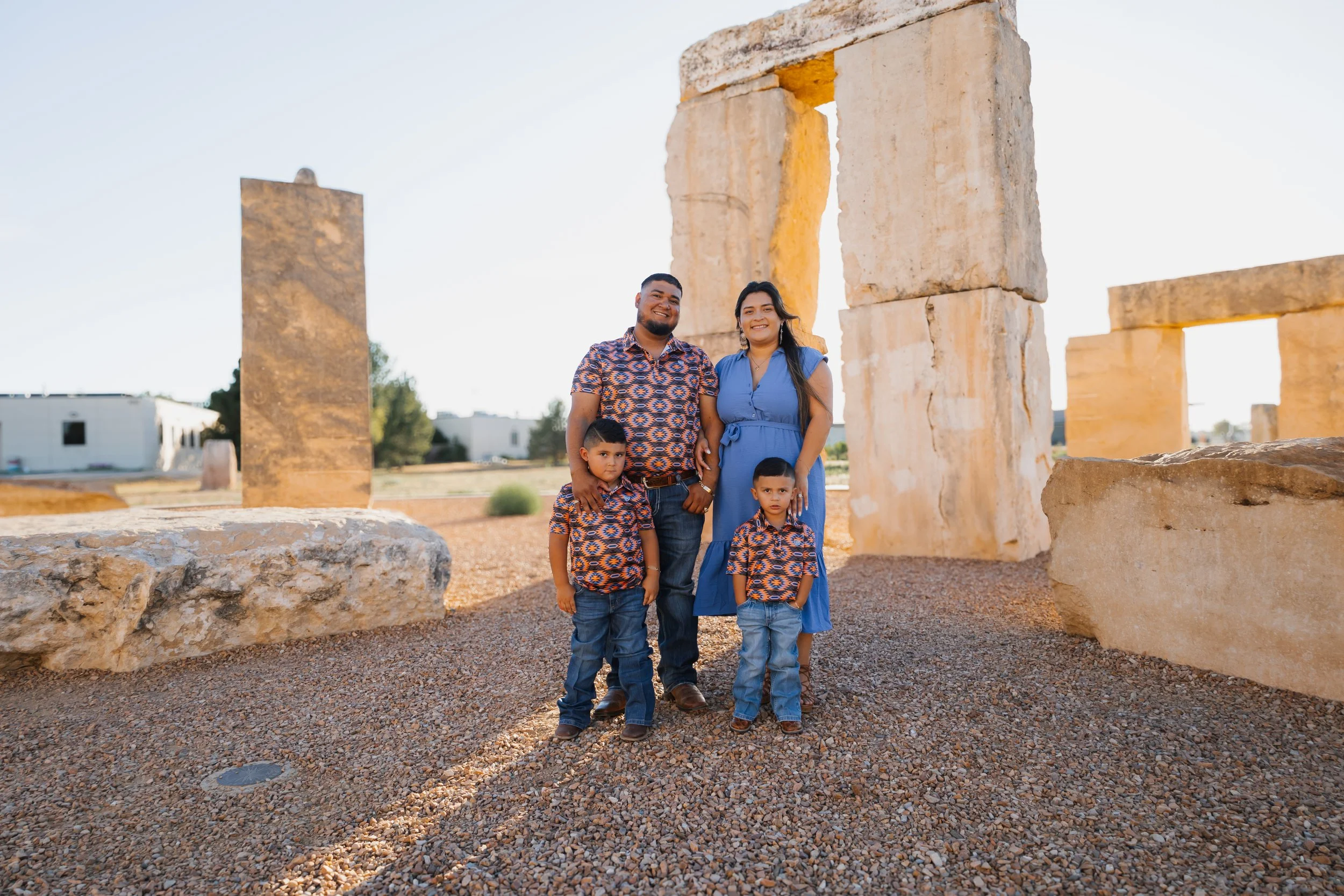 A family of five standing outdoors in front of ancient stone ruins, smiling at the camera, with sunlight casting long shadows on the ground.