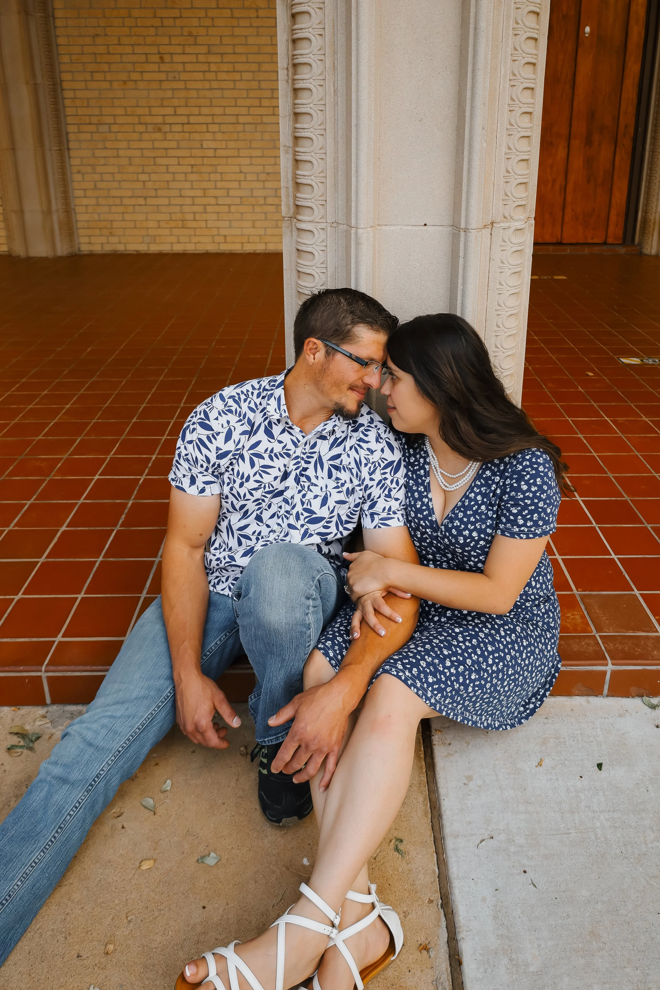 A man and woman sitting on the ground, leaning against a pillar, touching foreheads, with closed eyes in an intimate moment.