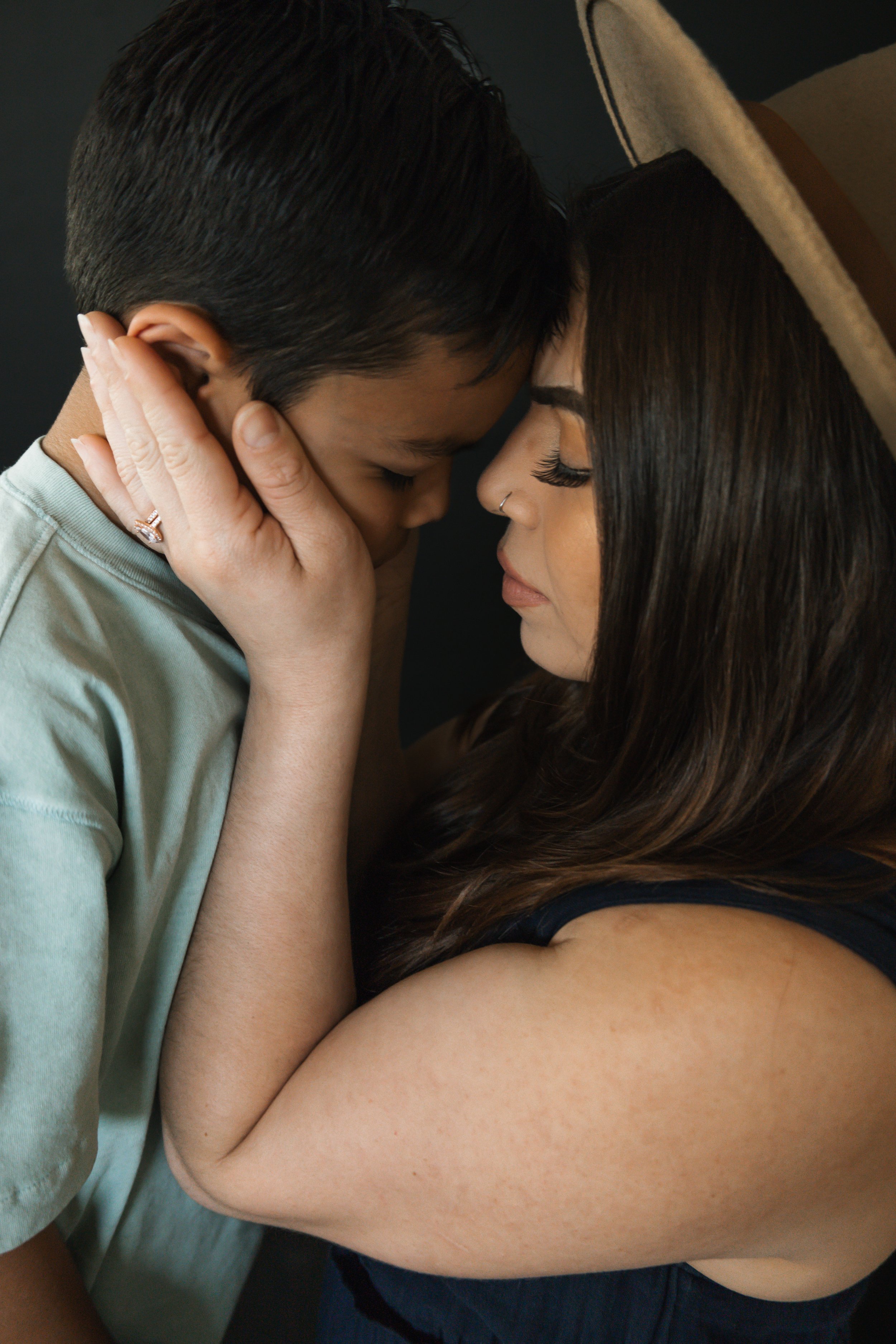 A close-up photo of a woman and a young boy with their foreheads touching, eyes closed, sharing an intimate moment. The woman has long dark hair, a nose piercing, wearing a hat, and a black sleeveless top. The boy has short dark hair, light skin, wearing a light-colored shirt.