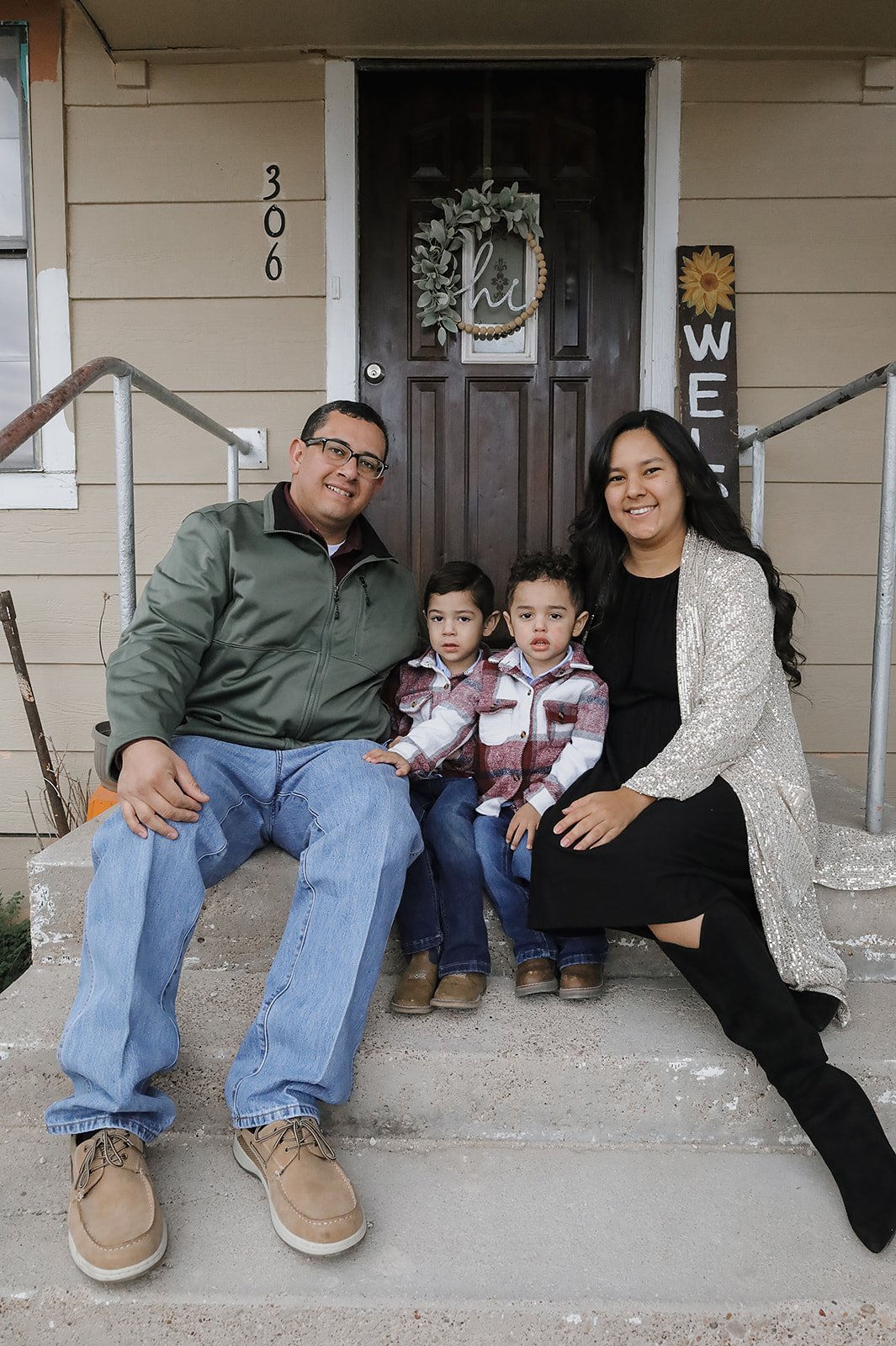 A family of four sitting on the front steps of a house, with a man, woman, and two young boys, smiling at the camera. The house has a dark wooden door with a decorative wreath and the number 306 on the wall beside it.