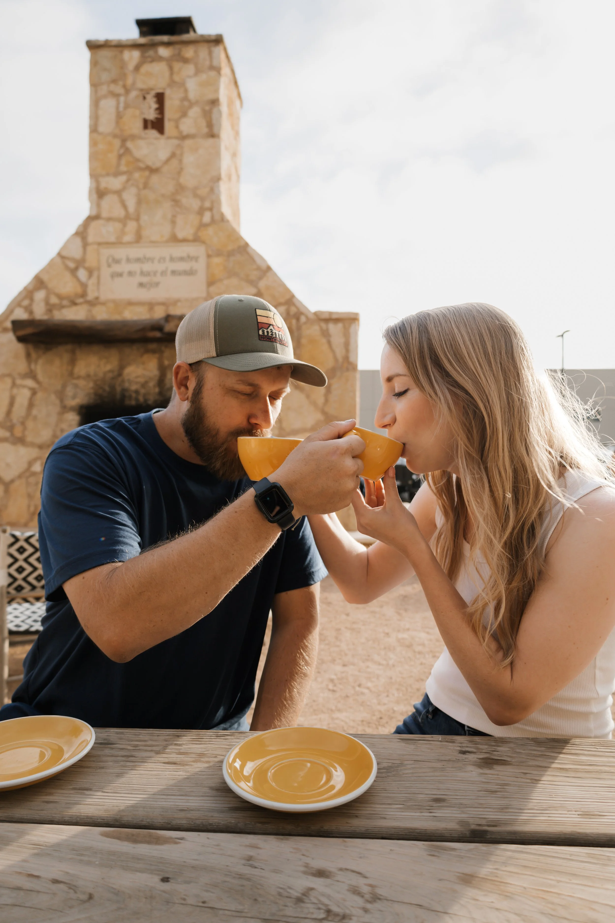 A man and woman sharing a drink outdoors at a wooden table, with a stone tower in the background.