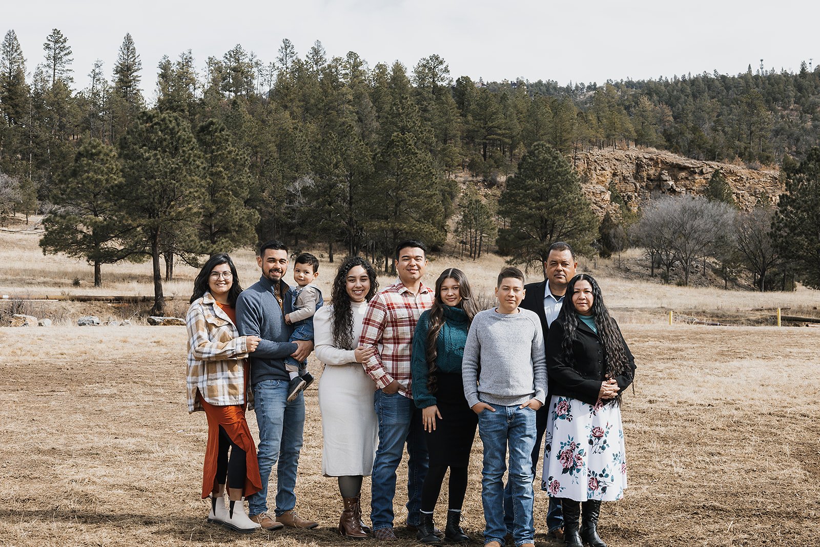 Family of nine standing outdoors in a field with trees and hills in the background.