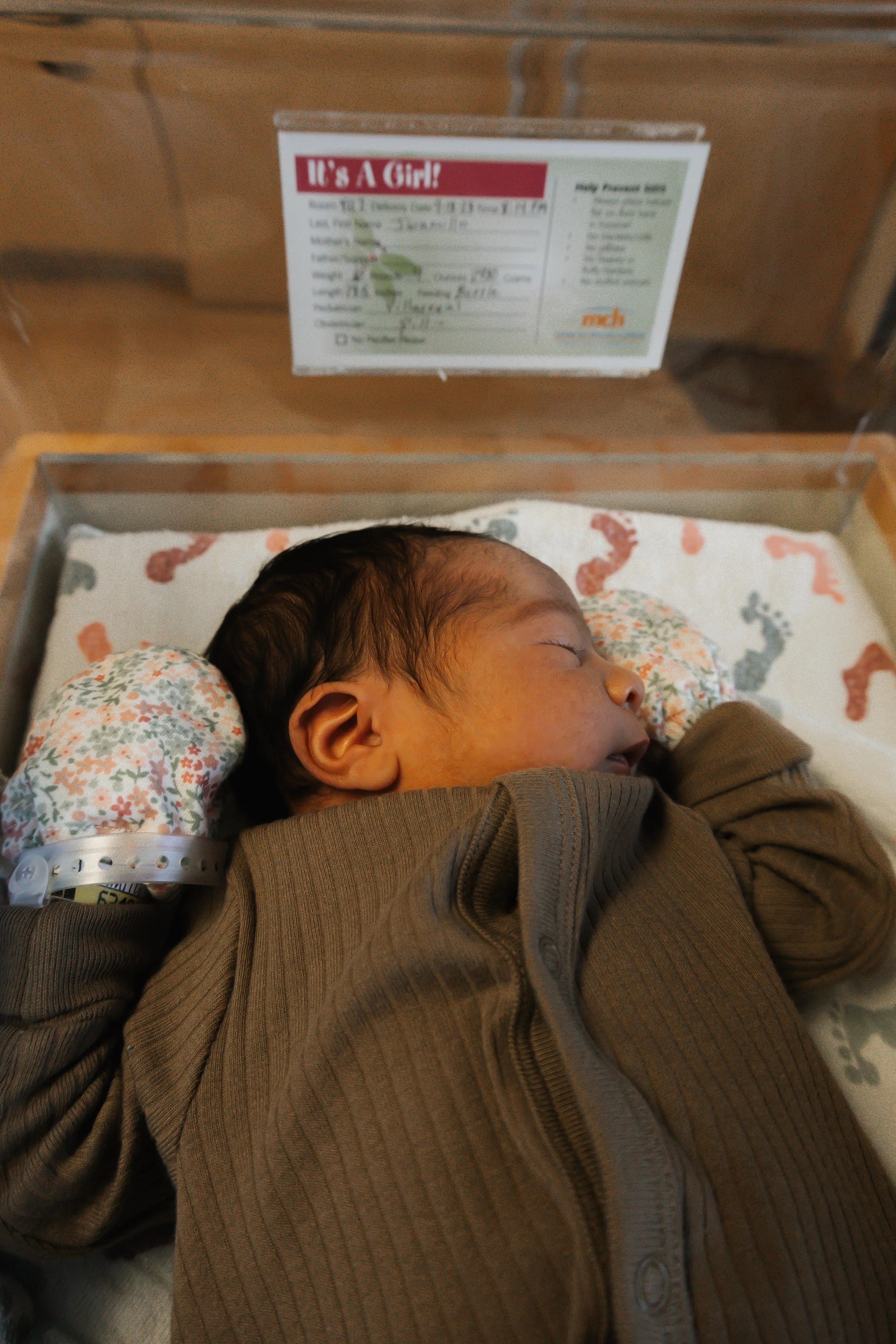 A newborn baby sleeping in a hospital bassinet, wearing a brown jacket, with a prenatal bracelet on its wrist. The bassinet has a white sheet with colorful footprints, and a pillow with a floral pattern. Behind, a card indicates the baby's gender as girl and contains birth details.