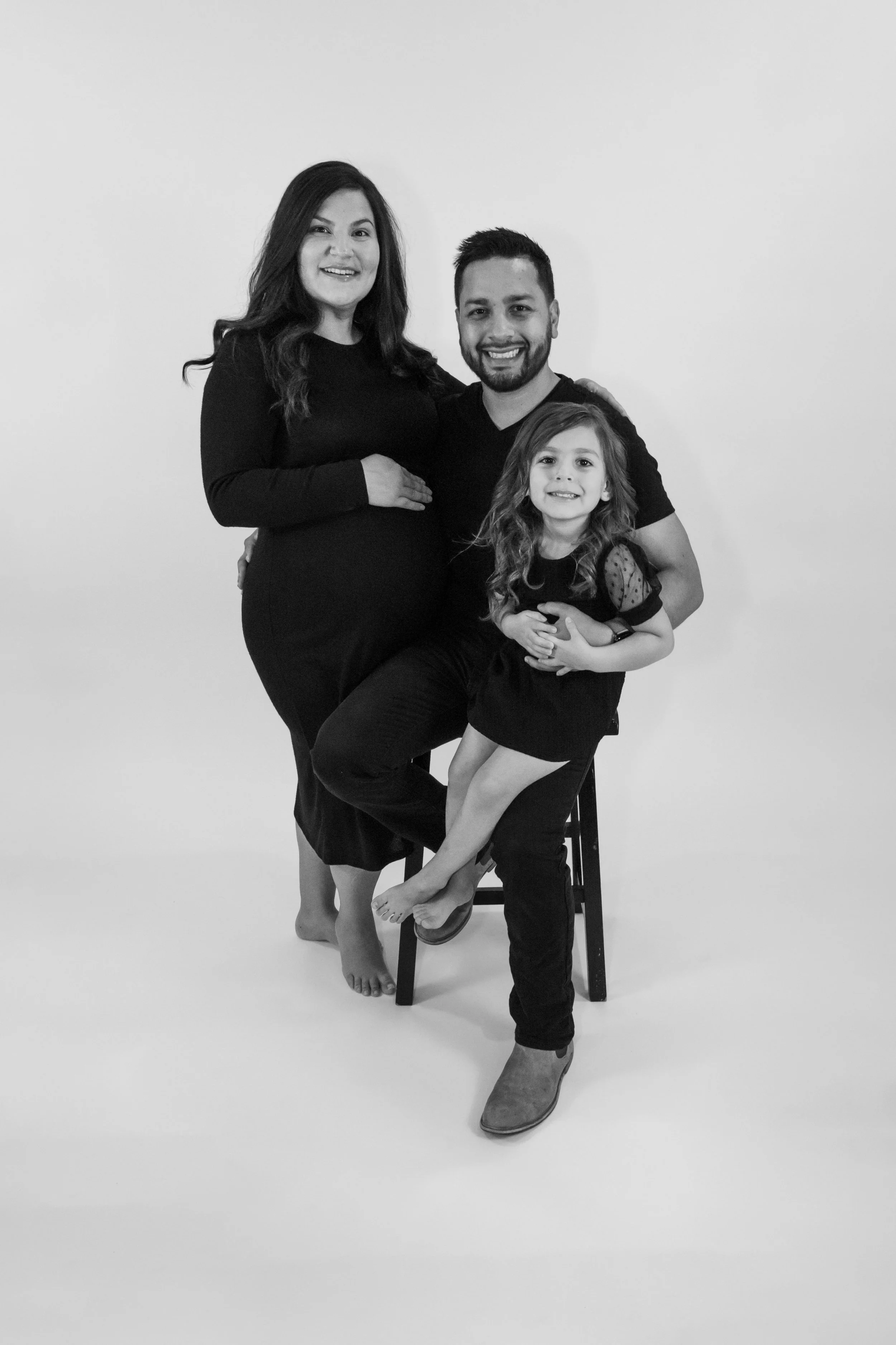 A black and white family portrait featuring a pregnant woman, a man, and a young girl, all smiling, with the girl sitting on his lap and the woman standing behind them.