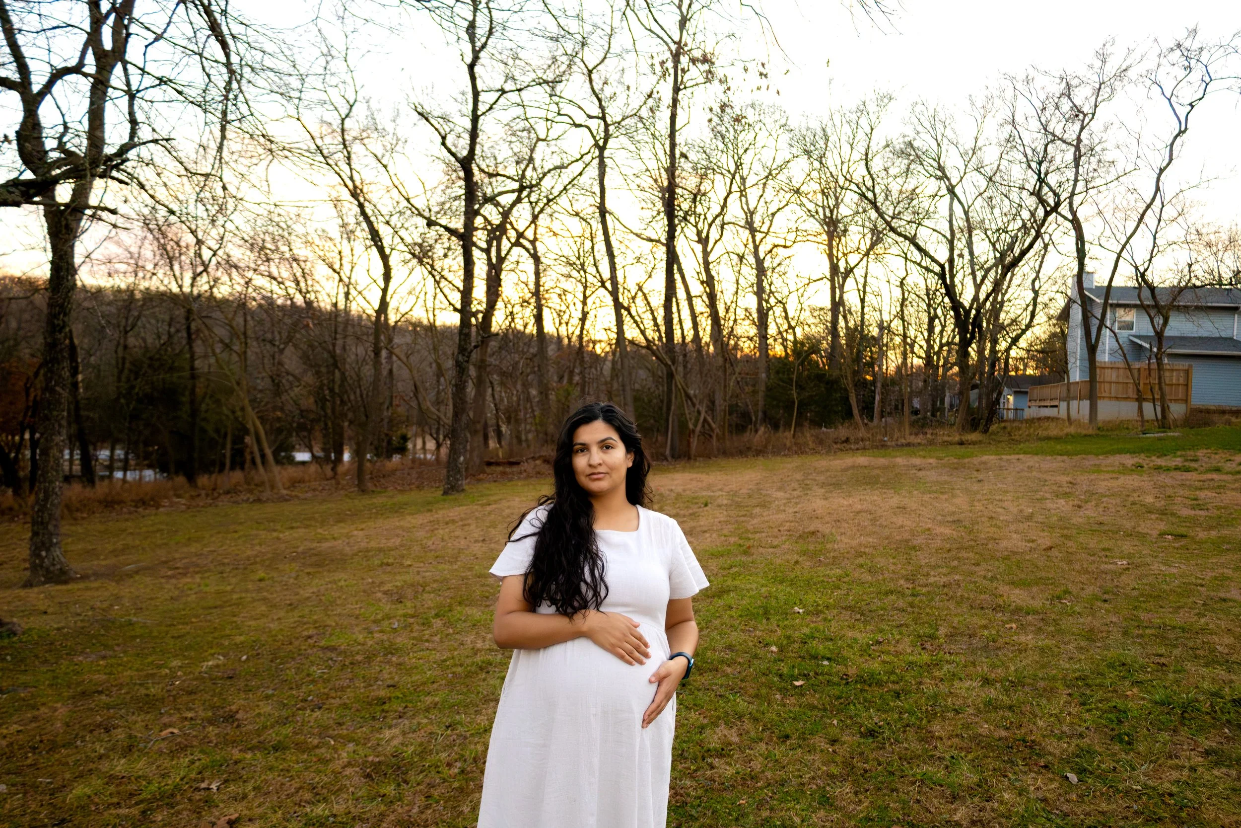 A pregnant woman in a white dress standing outdoors on a grassy lawn during sunset with leafless trees and houses in the background.