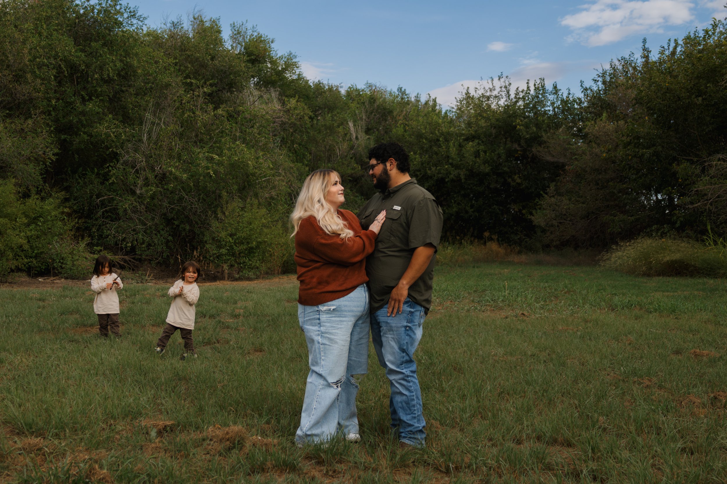 A couple stands close together in a grassy field, gazing into each other's eyes, with two children in the background playing among trees.
