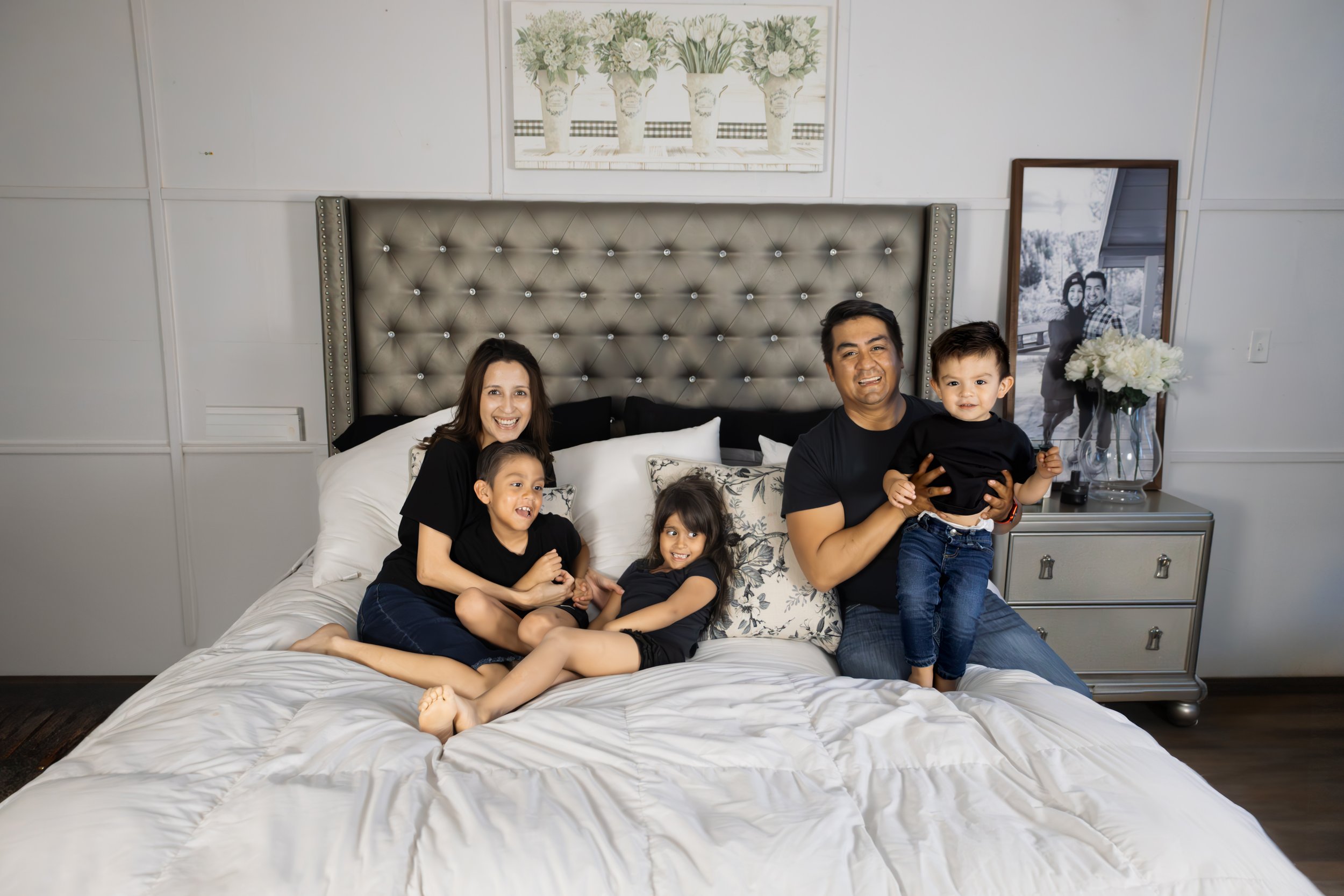 Family of five sitting on a bed in a bedroom, smiling at the camera. The mother is on the left, with three children in the middle, and the father is on the right holding a young boy. The room has a tufted headboard, floral artwork above, and a nightstand with a vase of white flowers and a framed black-and-white photo.