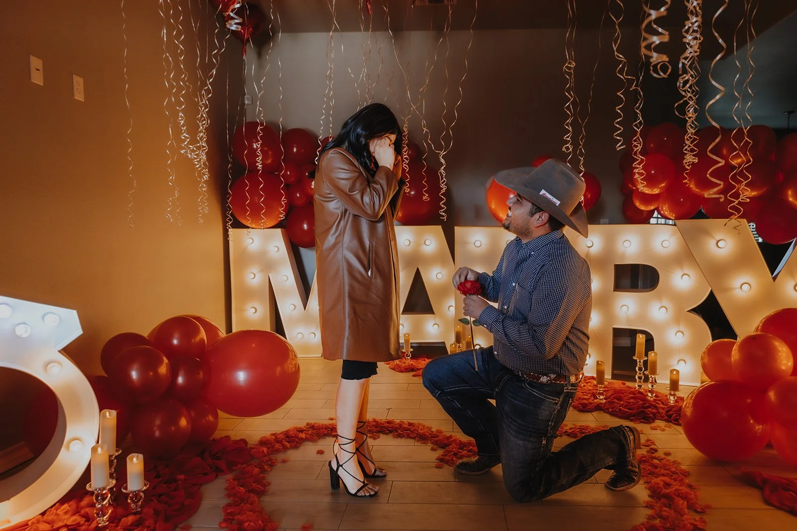 A man proposes to a woman during a romantic celebration with red balloons, candles, and big illuminated letters spelling 'MARRY' in the background. The woman looks surprised as the man kneels holding a red rose.
