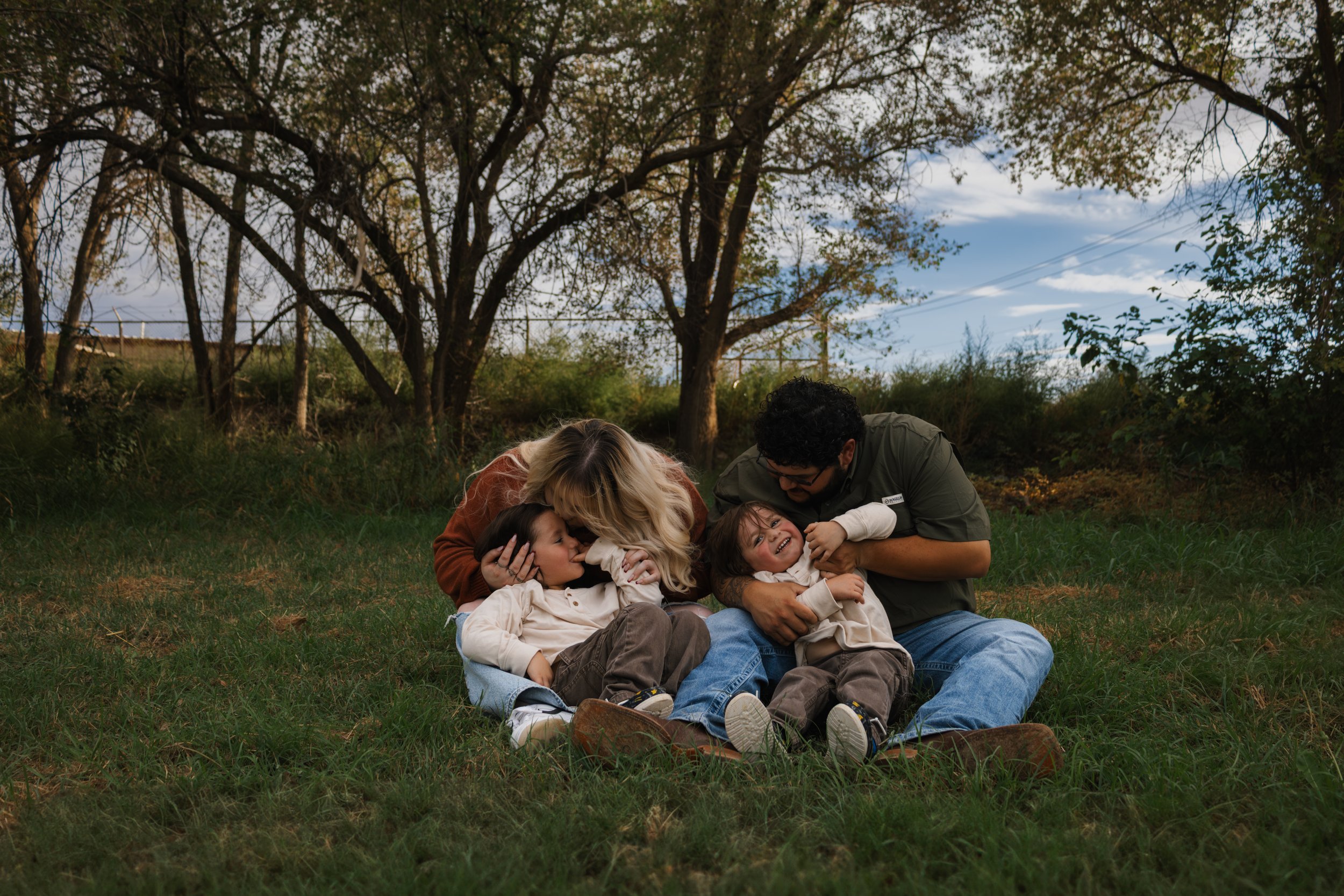 A family of four sitting on grass outdoors, playing and laughing together under trees during daytime.