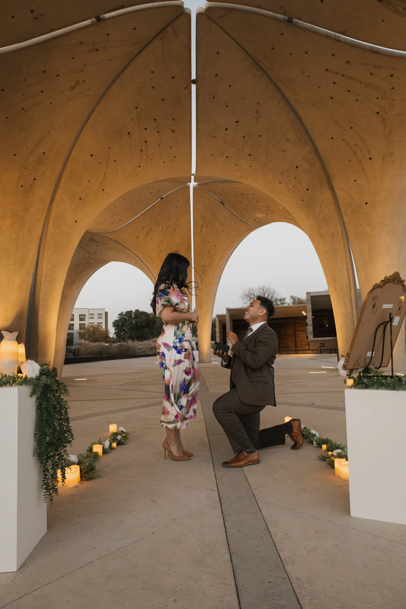 A man proposing to a woman at an outdoor wedding or proposal ceremony. The man is on one knee holding a box, likely with a ring, while the woman stands in front of him. They are under a decorative wooden arch with candles and greenery around.