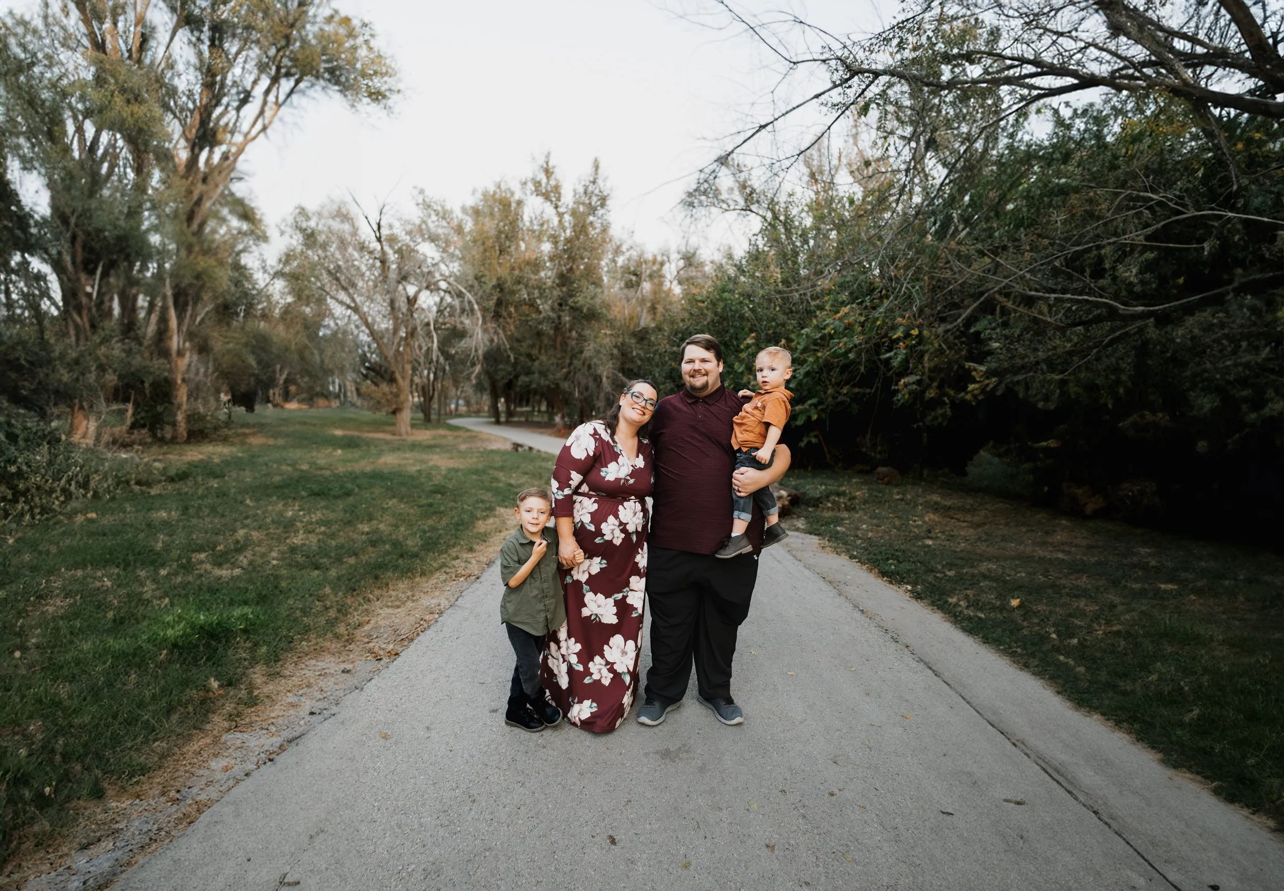 A family of four stand together on a park pathway surrounded by trees, with the parents smiling and holding their children, a boy and a toddler, during a fall season.