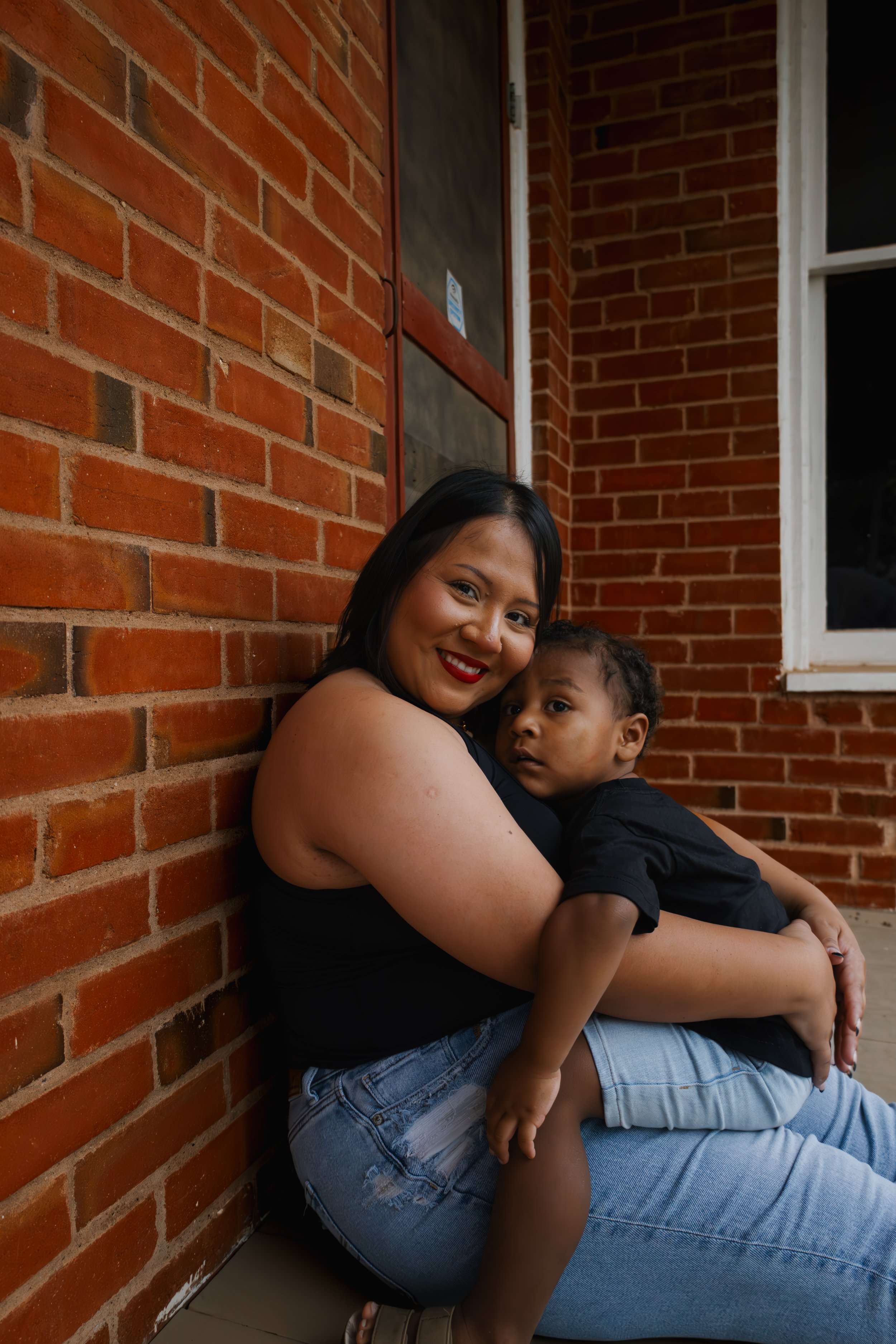 A woman with black hair and red lipstick sitting against a brick wall, smiling while holding a young boy with curly hair, dressed in black, who is sitting on her lap.