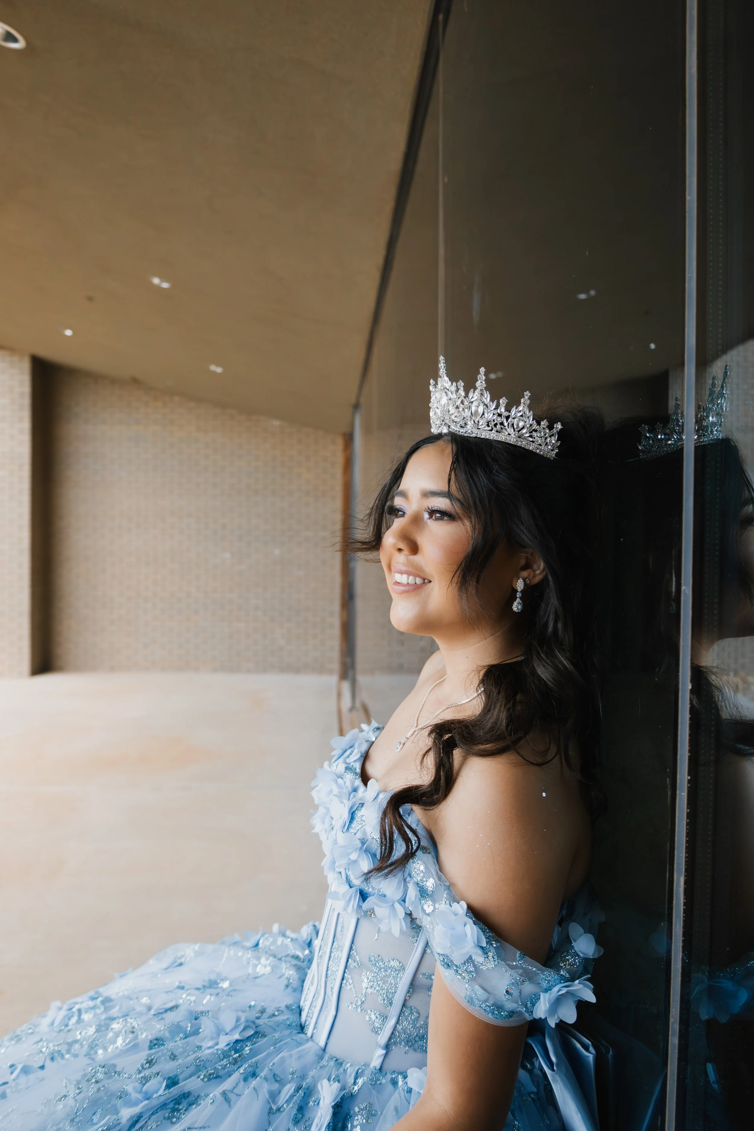Young woman wearing a silver crown and a blue off-shoulder dress, smiling and sitting against a wall.