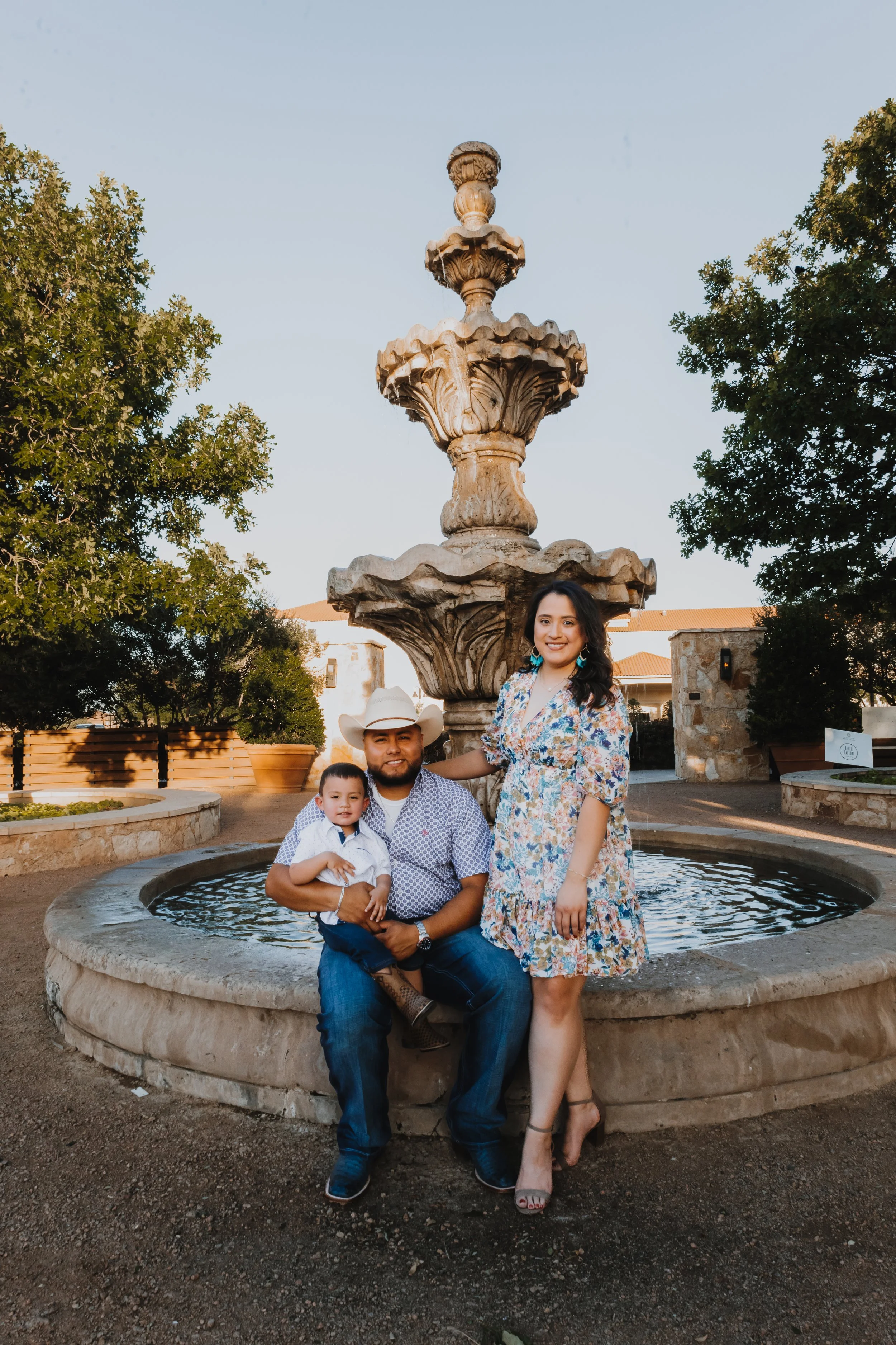 A family of three posing in front of a decorative stone fountain with water, surrounded by trees and a clear sky.