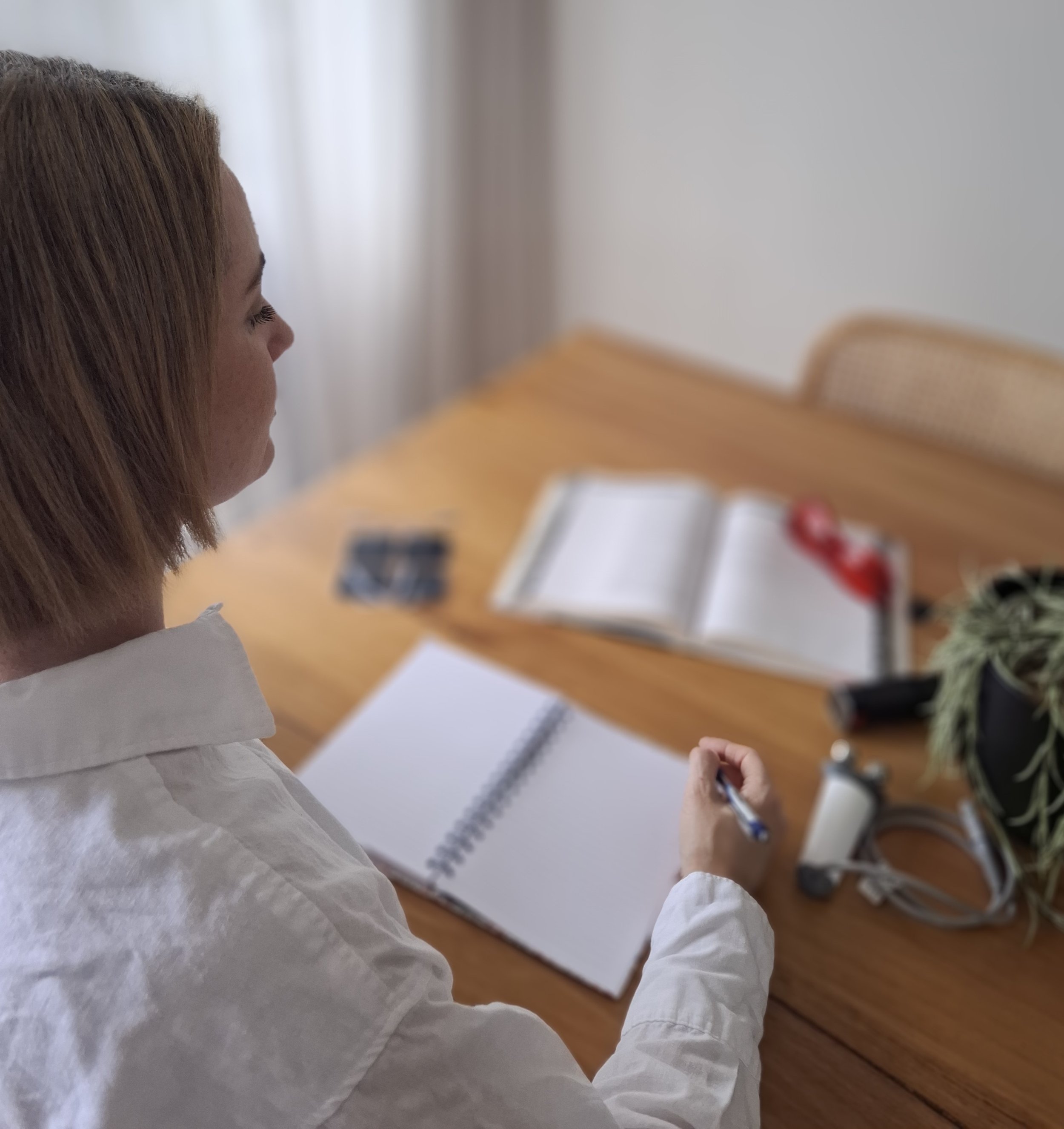 A woman with shoulder-length hair wearing a white shirt, sitting at a wooden table with an open notebook, a pen, and various items including a plant, a blood pressure monitor, an open book, and other objects on the table.